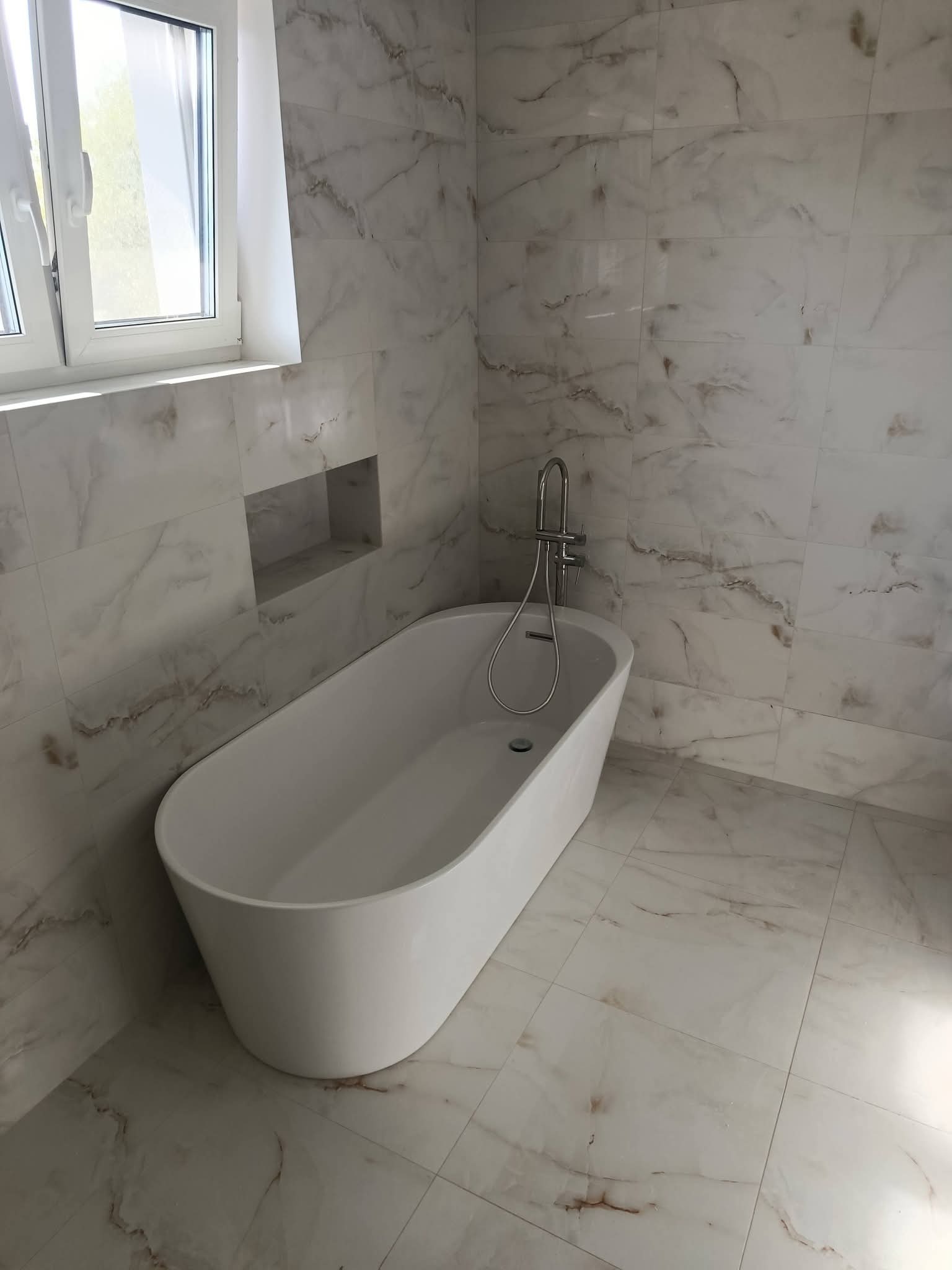 Nowoczesna łazienka z wolnostojącą białą wanną i marmurowymi płytkami. Wnęka w ścianie dodaje charakteru. Minimalistyczna armatura.