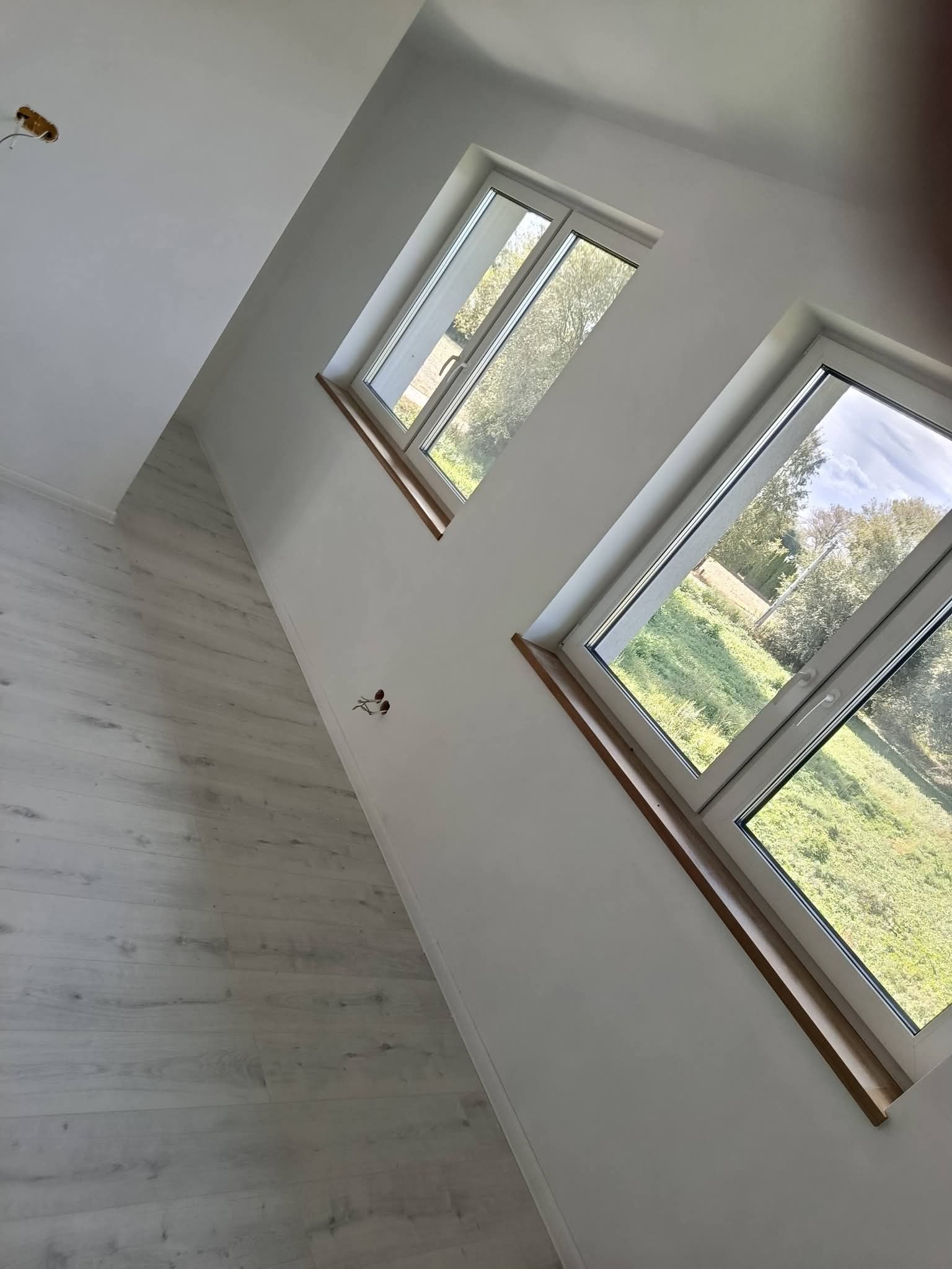 Jasne panele podłogowe i dwa okna z drewnianymi parapetami w nowym wnętrzu w Warszawie. Świeżo pomalowane ściany i widok na zieleń za oknem.