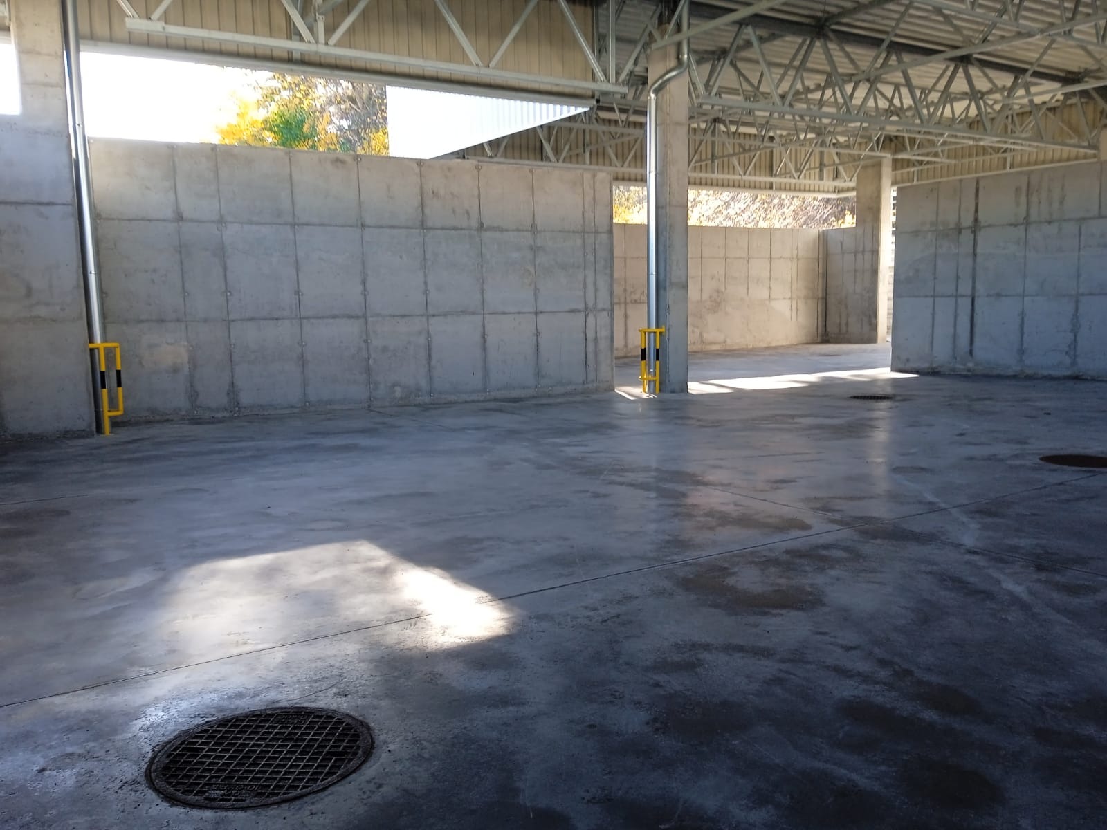 Wnętrze hali z betonowymi ścianami działowymi i podłogą, metalowa konstrukcja dachu, słupy z żółto-czarnym zabezpieczeniem, widoczne studzienki kanalizacyjne.