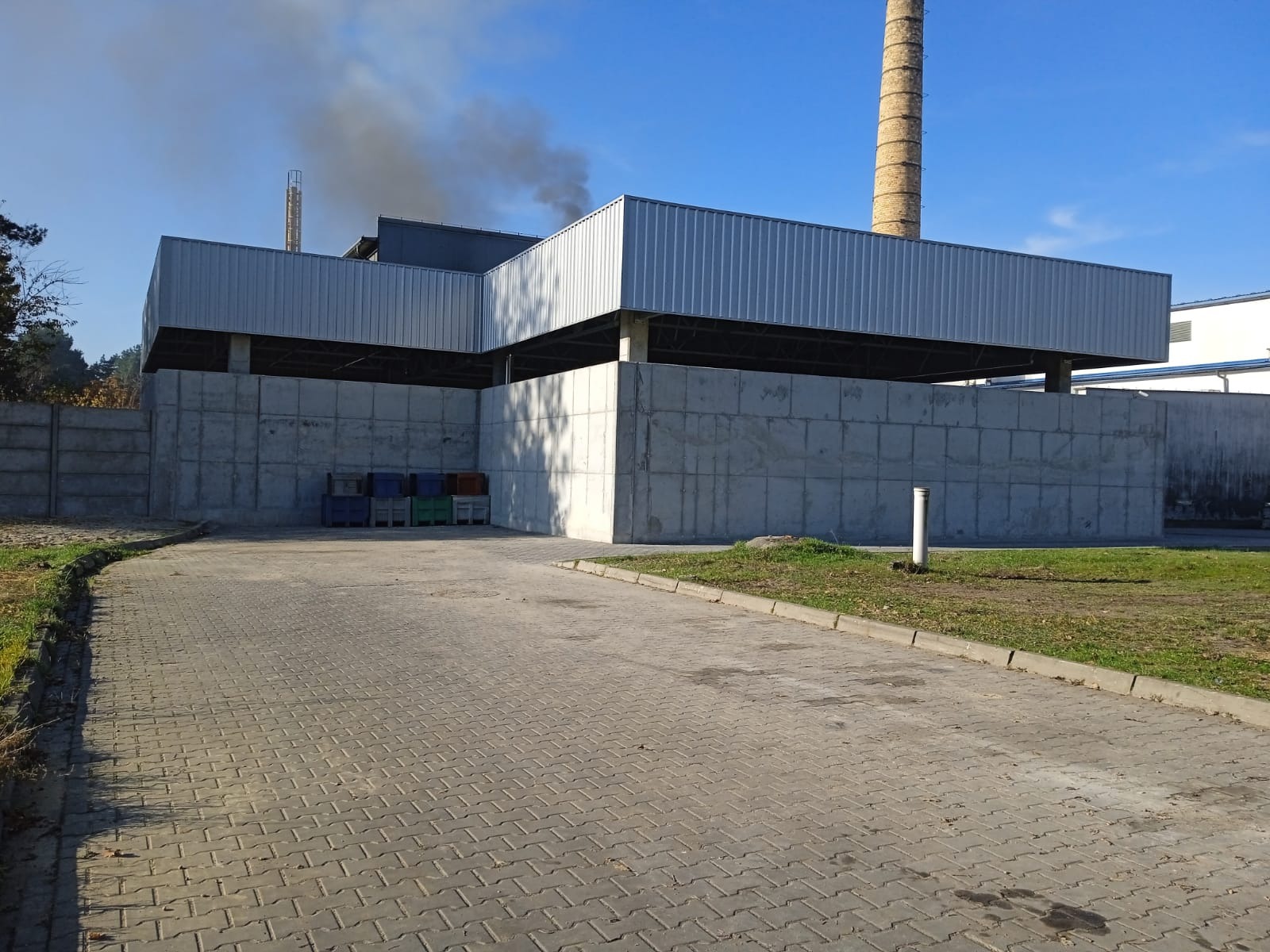 Żelbetowa konstrukcja przemysłowa z widocznym kominem w tle i paletami przy ścianie, sfotografowana w słoneczny dzień.