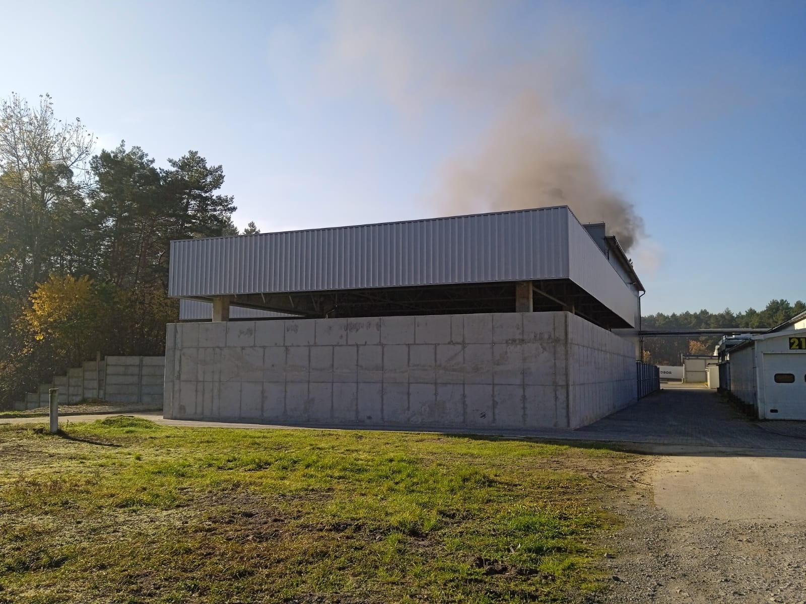 Wznoszony budynek przemysłowy o konstrukcji żelbetowej i metalowym dachu, z widocznym dymem wydobywającym się z wnętrza, otoczony zielenią i drzewami w słoneczny dzień.