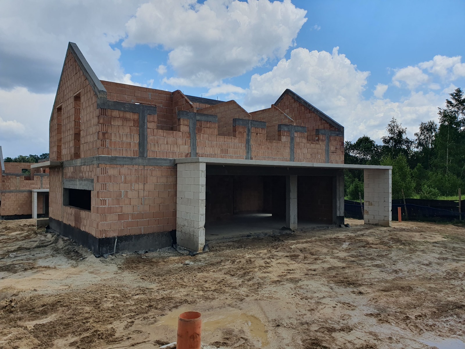 Surowy stan domu murowanego z betonu komórkowego, widoczne ściany nośne, otwory okienne i drzwiowe, zbrojenia, oraz teren budowy pokryty piaskiem i błotem, błękitne niebo z chmurami w tle.