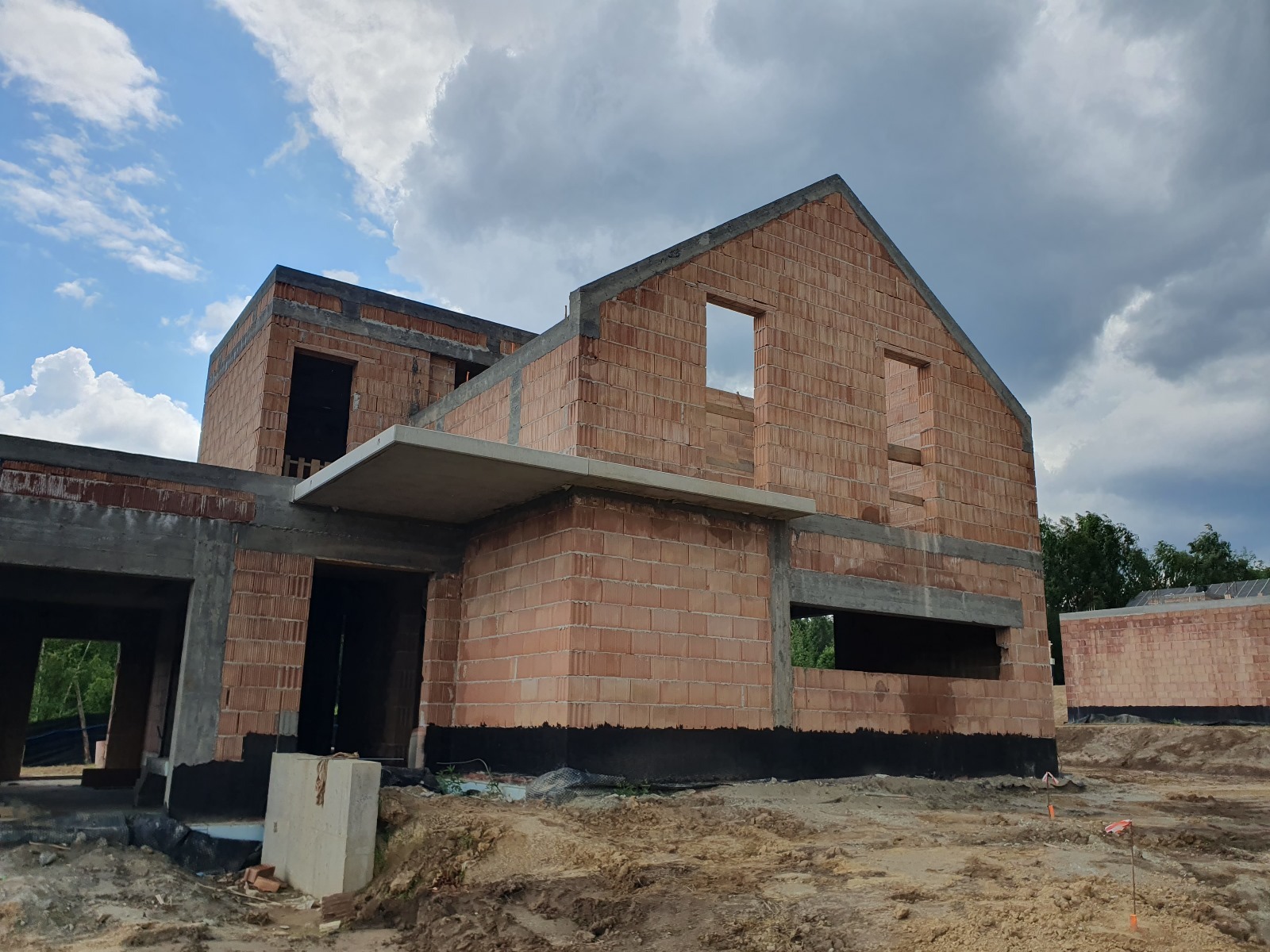 Surowy stan dwukondygnacyjnego domu z czerwonej cegły, z widocznymi otworami okiennymi i betonowymi nadprożami, na tle pochmurnego nieba.