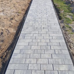 Embruk - Ścieżka z szarej kostki brukowej ułożona wzdłuż trawnika, widok z perspektywy. Prace brukarskie w toku w Białej Podlaskiej. Widoczny stos kostki i pracownicy.