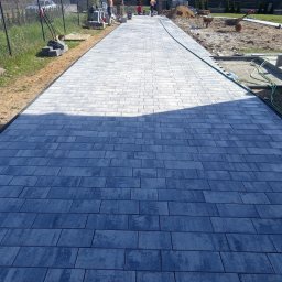 Embruk - Długa, szara ścieżka z kostki brukowej prowadzi przez posesję. Widoczne narzędzia i materiały budowlane, sugerujące trwające prace wykończeniowe.