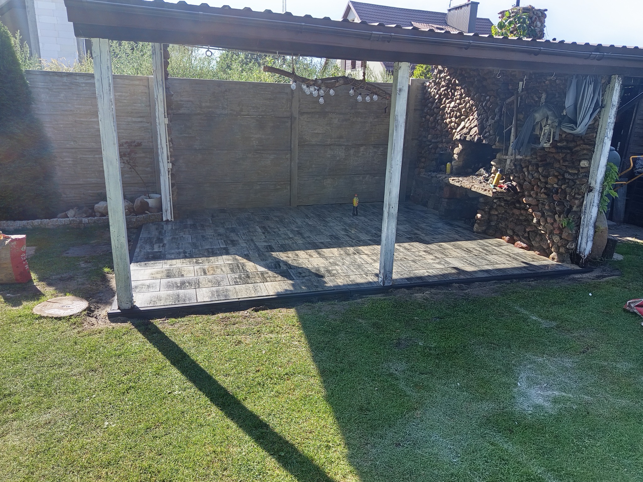 Zadaszona altana z kostki brukowej w szaro-brązowej tonacji, obok trawnik. Fragment dekoracyjnego muru z kamienia. Elementy oświetlenia i dekoracje ogrodowe.