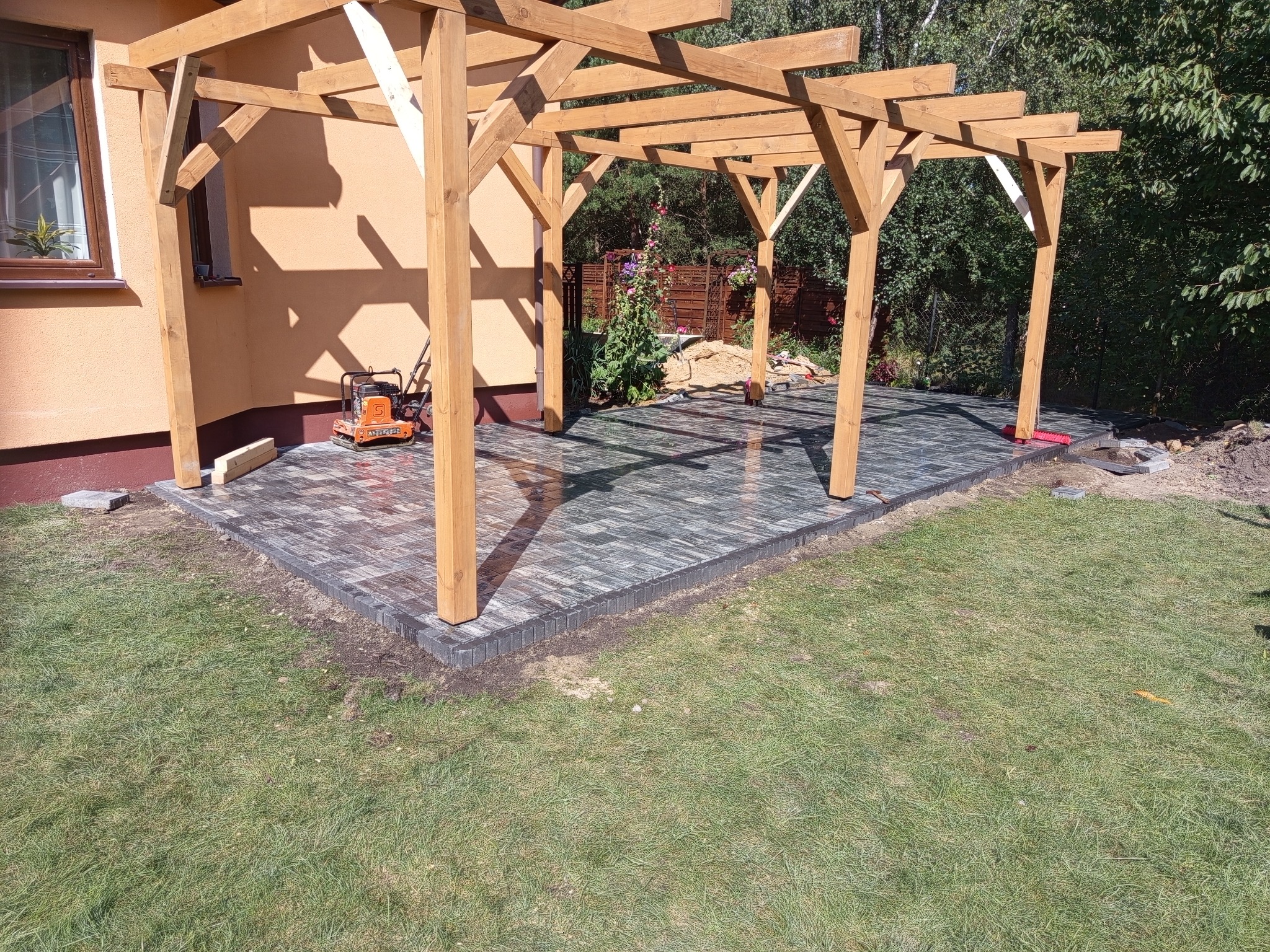 Nowoczesna, szara kostka brukowa pod drewnianą pergolą, z zagęszczarką obok. Estetyczne wykończenie tarasu w ogrodzie z zielonym trawnikiem w słoneczny dzień.