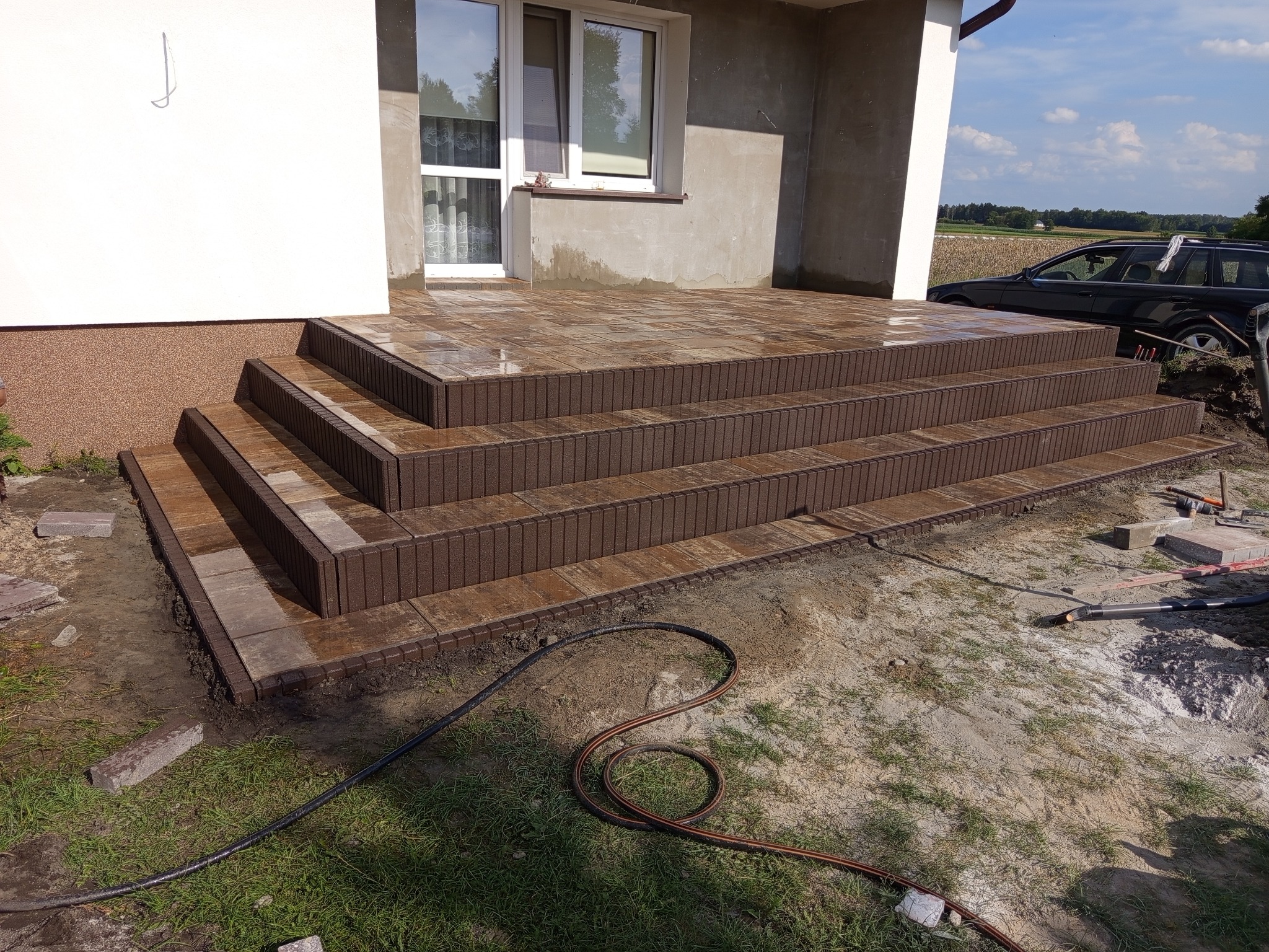 Schody wejściowe z kostki brukowej w odcieniach brązu, prowadzące do domu. Widoczne narzędzia i elementy otoczenia w trakcie prac wykończeniowych.