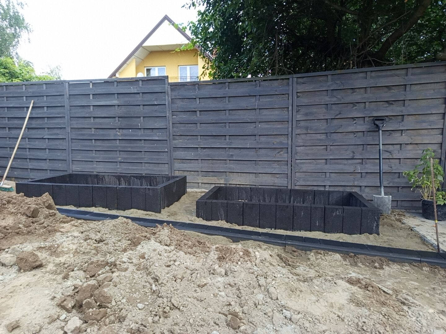 Przygotowane podłoże pod rabaty z obrzeżami z kostki, na piasku, z widoczną łopatą i drewnianym płotem w tle, w trakcie prac ogrodowych.