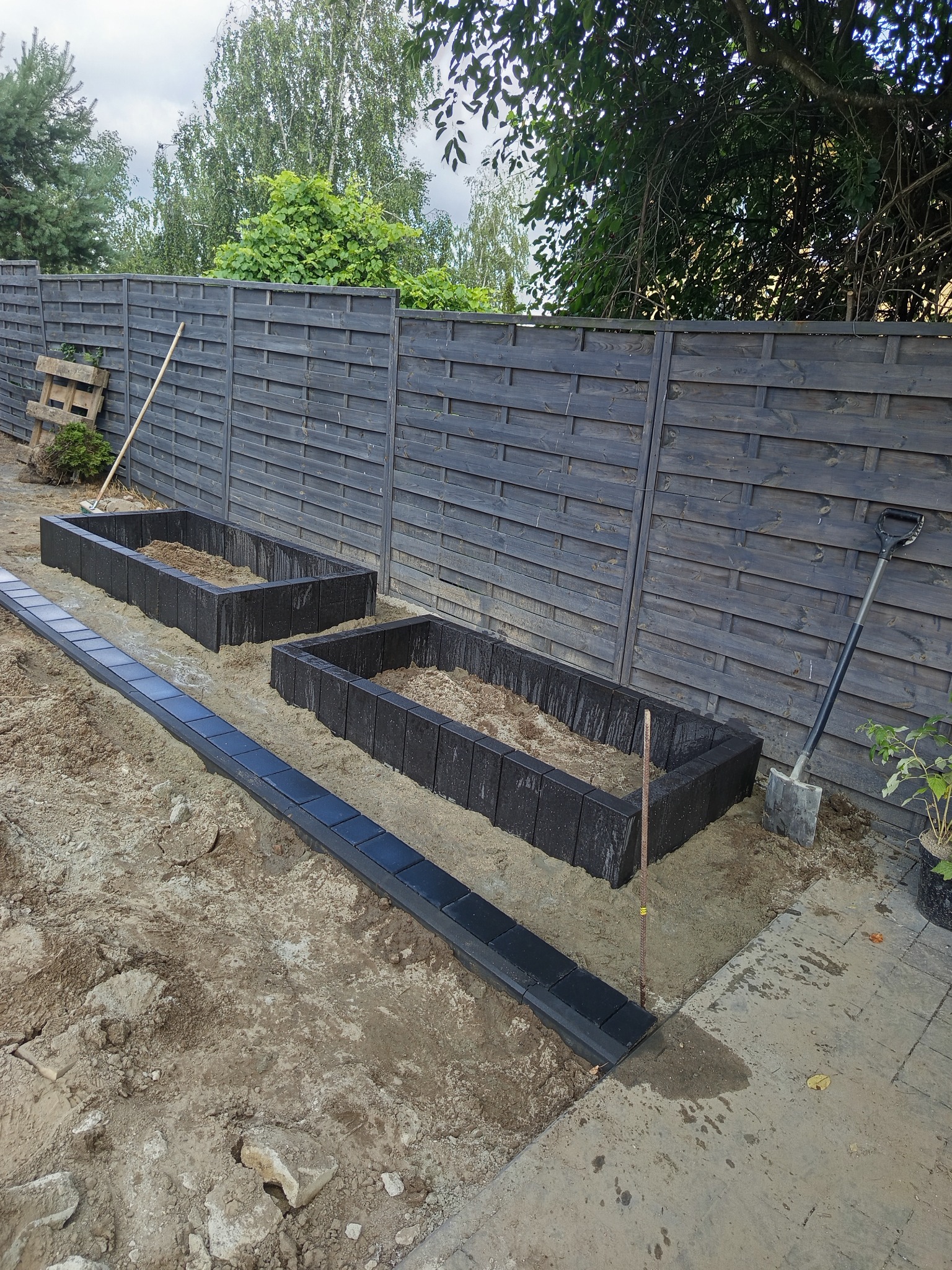 Przygotowanie rabat z kostki brukowej w ogrodzie, widoczne obrzeża i wypełnione ziemią koryta, szpadel oparty o drewniane ogrodzenie.