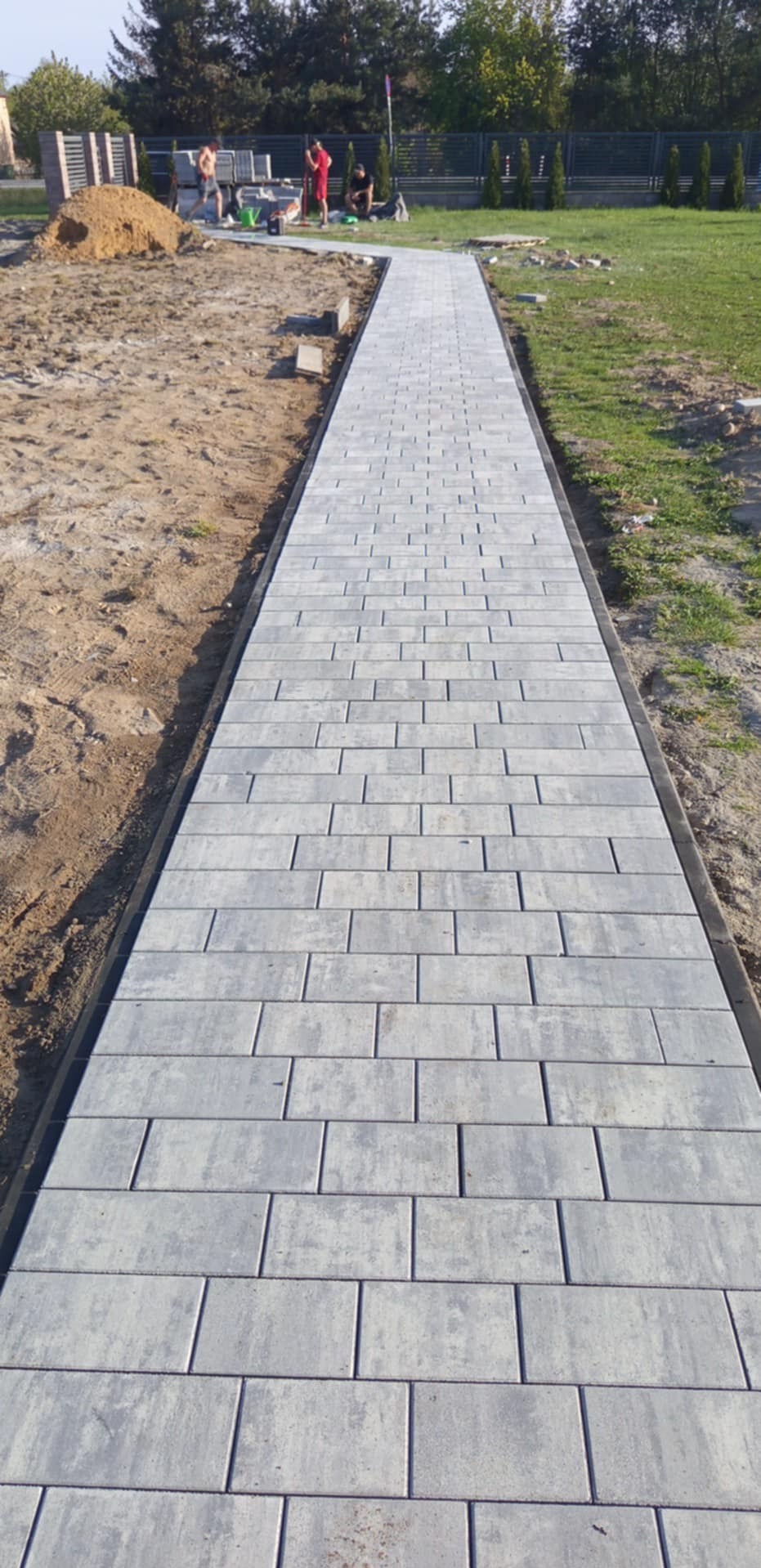 Ścieżka z szarej kostki brukowej ułożona wzdłuż trawnika, widok z perspektywy. Prace brukarskie w toku w Białej Podlaskiej. Widoczny stos kostki i pracownicy.