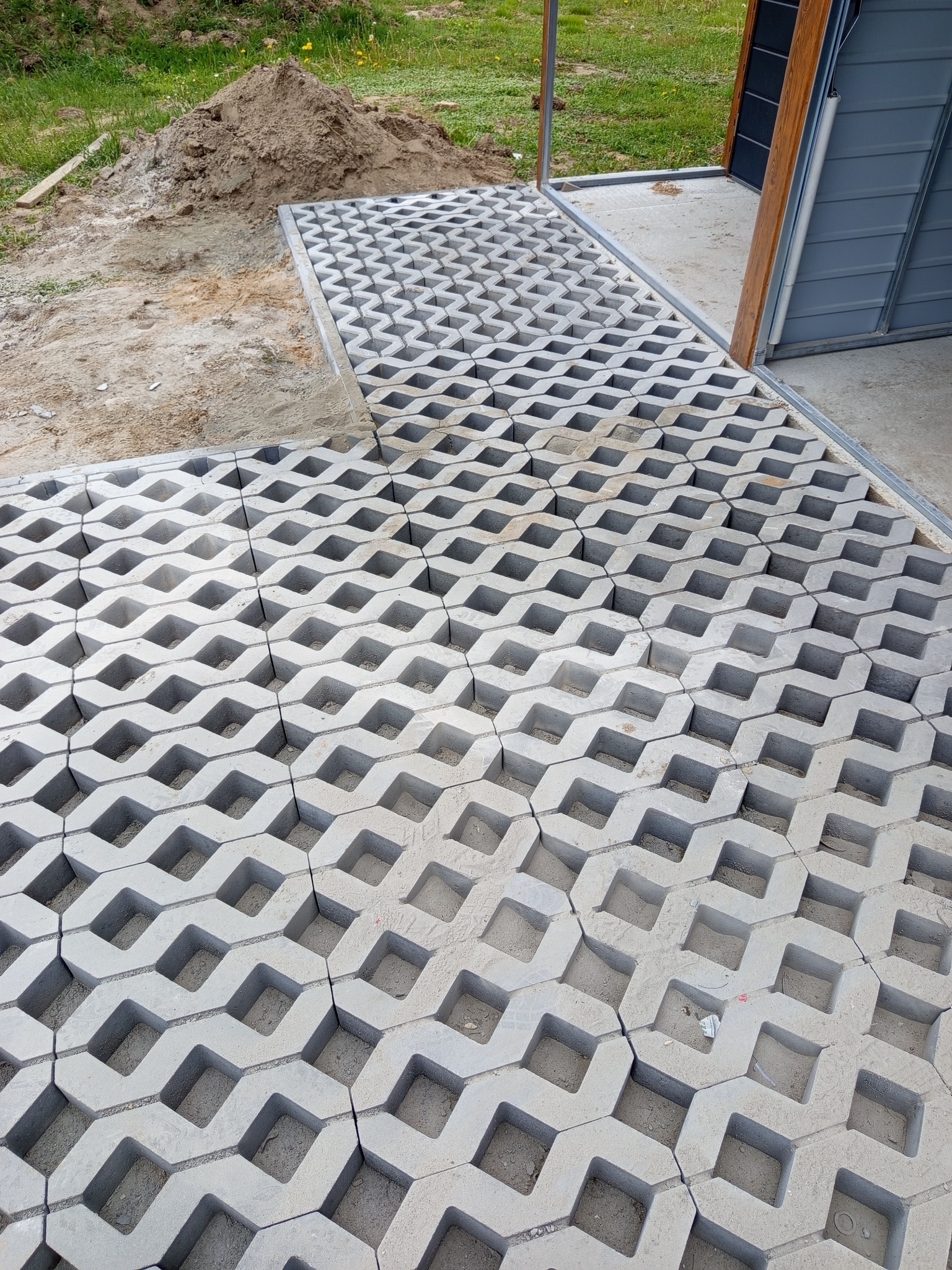 Szara, ażurowa kostka brukowa ułożona przy wjeździe do garażu. Częściowo widoczna sterta ziemi. Widok z góry.