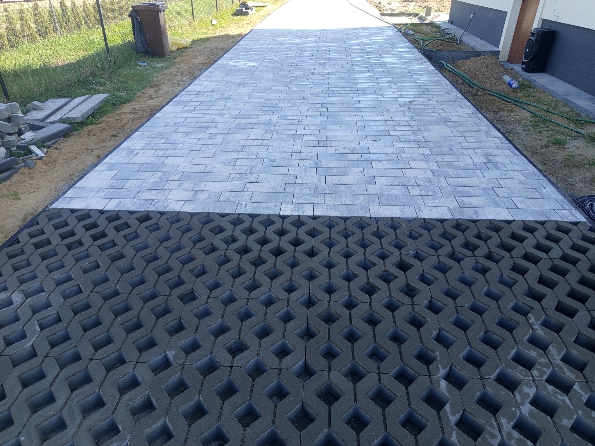 Ścieżka z jasnoszarej kostki brukowej ułożona na czarnych kratkach trawnikowych. Widoczne przygotowane obrzeża i zapas kostki.