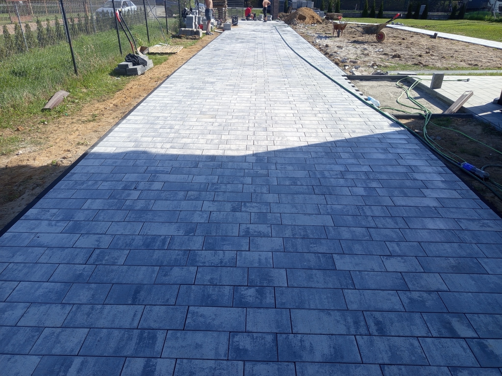 Długa, szara ścieżka z kostki brukowej prowadzi przez posesję. Widoczne narzędzia i materiały budowlane, sugerujące trwające prace wykończeniowe.