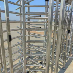 Metalowy, obrotowy system kontroli dostępu na zewnątrz budynku, widoczny na tle betonowej nawierzchni i błękitnego nieba.