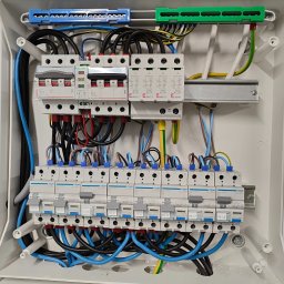 Piotr Jurczenko - Rozdzielnica elektryczna w białej obudowie, widoczne bezpieczniki, przewody w kolorach niebieskim, czarnym, brązowym, żółto-zielonym, listwy zaciskowe w kolorze niebieskim i zielonym, wykonana...