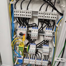 Piotr Jurczenko - Wnętrze rozdzielnicy elektrycznej z widocznymi bezpiecznikami, przewodami w kolorach niebieskim, żółto-zielonym i czarnym, oraz blokami zaciskowymi z oznaczeniami 'Zasilanie UPS' i 'Wyjście UPS'.