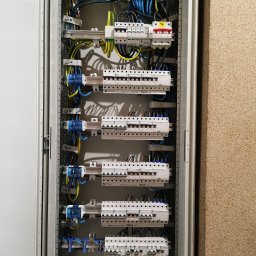 Piotr Jurczenko - Otwarta szafa rozdzielcza z bezpiecznikami, przewodami i listwami montażowymi, widok z bliska na instalację elektryczną wewnątrz pomieszczenia.