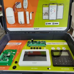 Zestaw demonstracyjny systemu alarmowego w otwartej walizce, prezentujący centralę, manipulatory, detektory i aplikację mobilną na smartfonach, marki Satel.