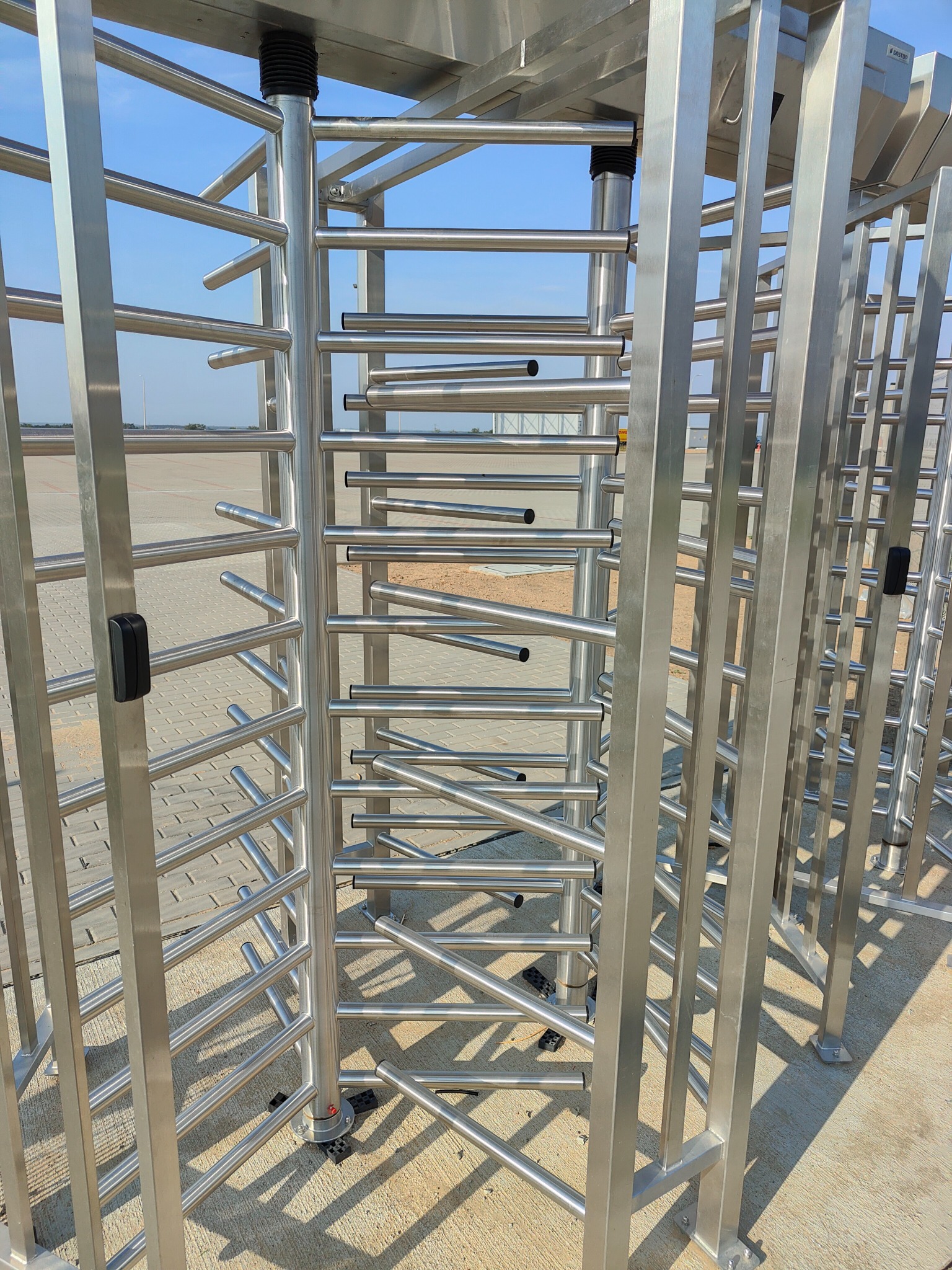 Metalowy, obrotowy system kontroli dostępu na zewnątrz budynku, widoczny na tle betonowej nawierzchni i błękitnego nieba.