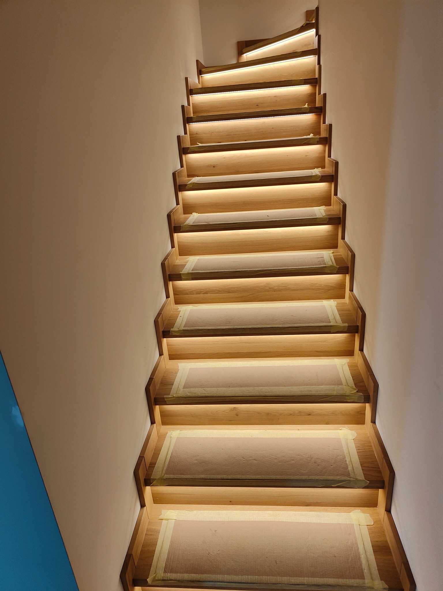 Drewniane schody z podświetleniem LED na każdym stopniu, zabezpieczone taśmą malarską podczas prac wykończeniowych.