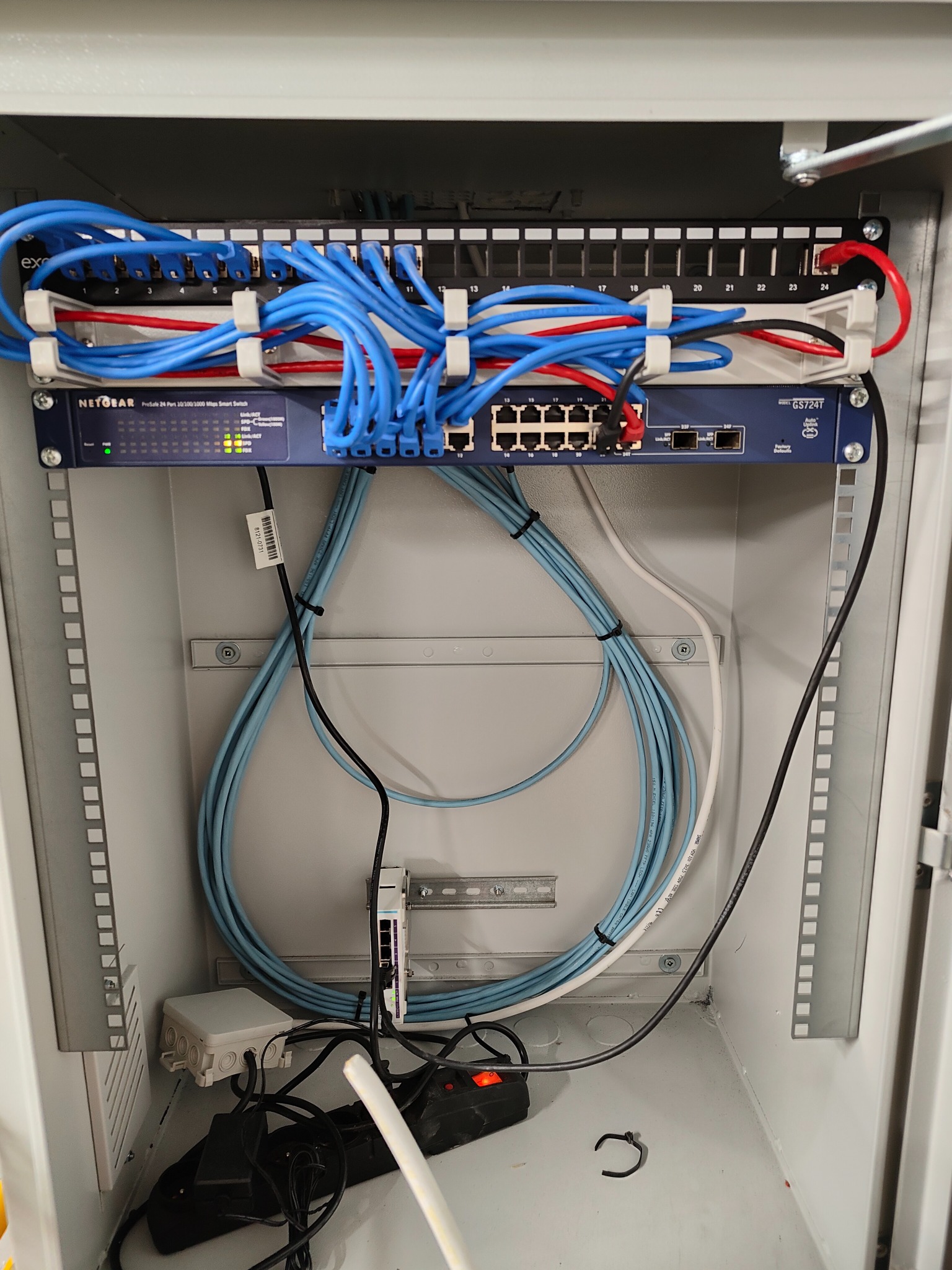 Szafa teleinformatyczna z zamontowanym switchem Netgear GS724T, patch panelem i okablowaniem strukturalnym w kolorach niebieskim, białym i czarnym, widoczne zasilacze i kable zasilające na dnie szafy.