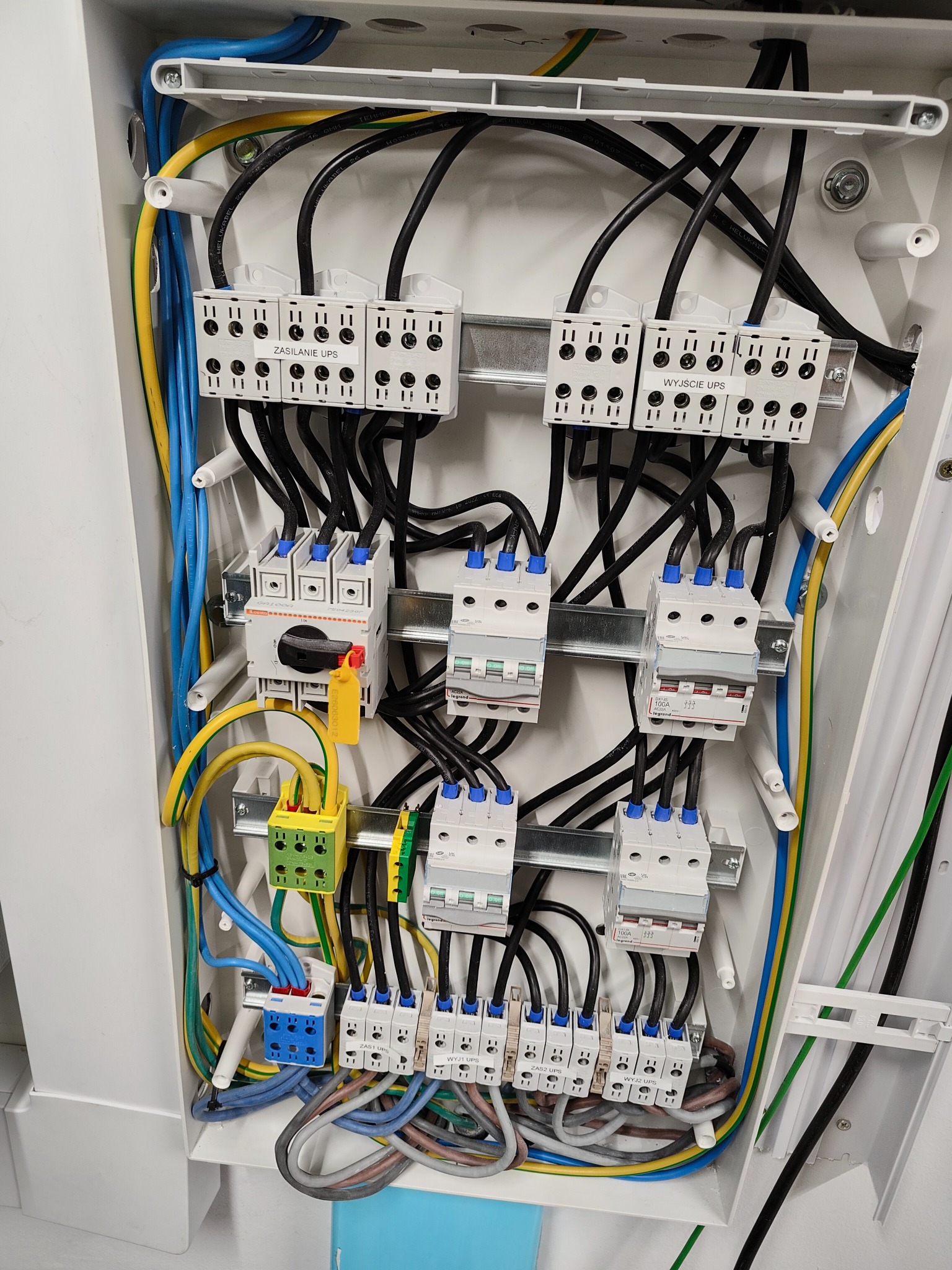 Wnętrze rozdzielnicy elektrycznej z widocznymi bezpiecznikami, przewodami w kolorach niebieskim, żółto-zielonym i czarnym, oraz blokami zaciskowymi z oznaczeniami 'Zasilanie UPS' i 'Wyjście UPS'.