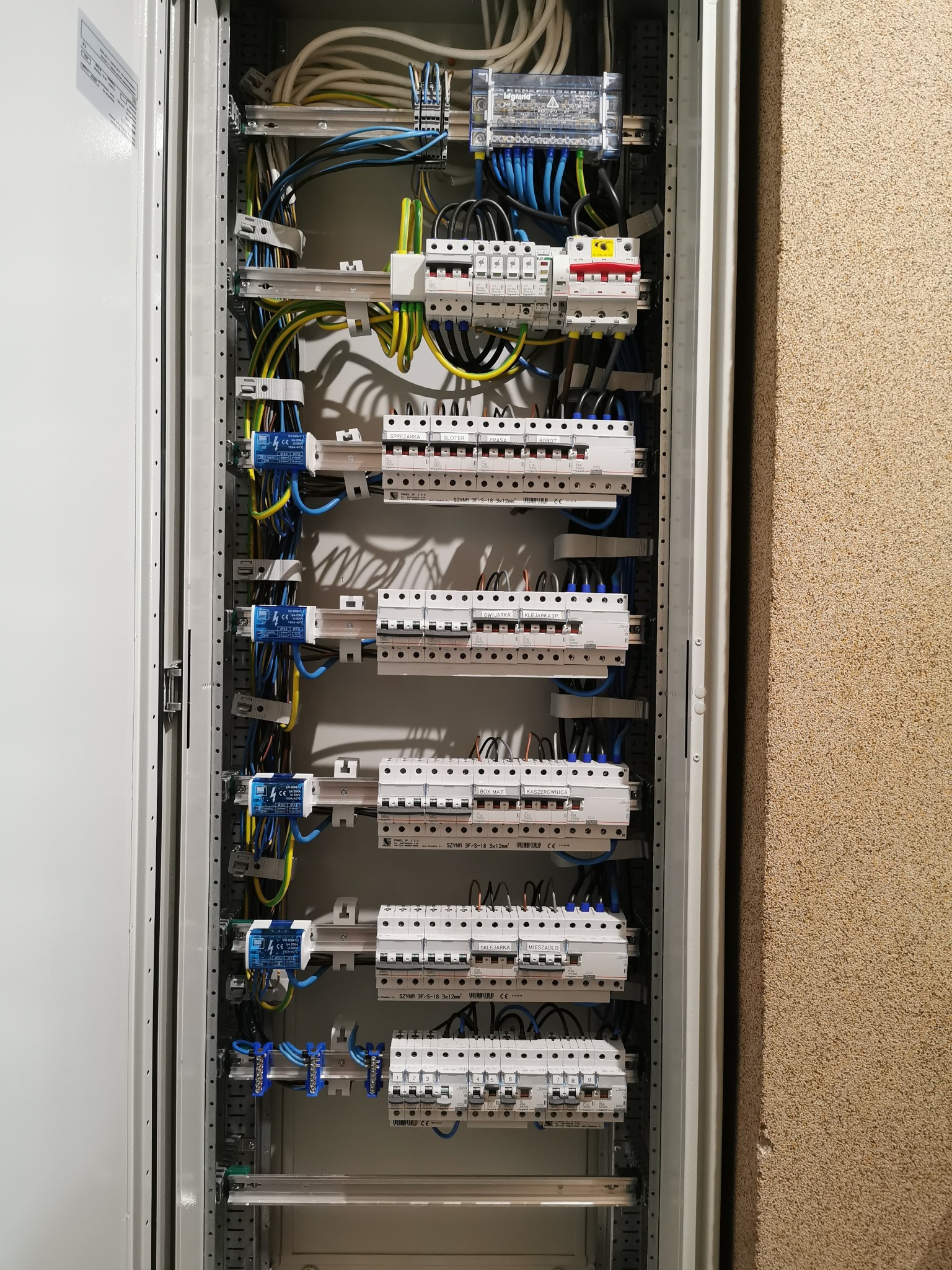 Otwarta szafa rozdzielcza z bezpiecznikami, przewodami i listwami montażowymi, widok z bliska na instalację elektryczną wewnątrz pomieszczenia.