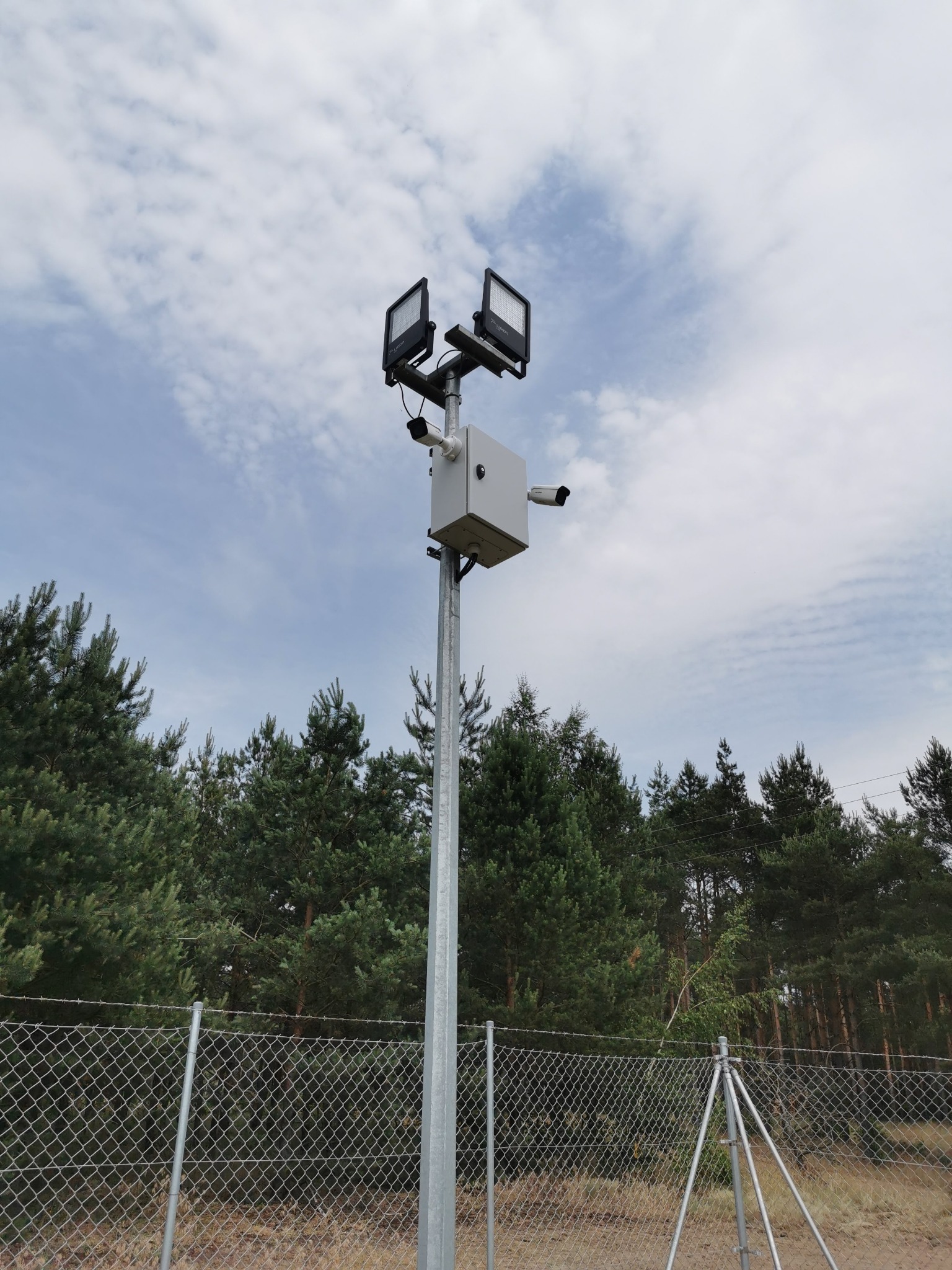 Słup oświetleniowo-monitoringowy z dwoma reflektorami LED i kamerami, umieszczony na obrzeżach lasu, widoczny płot z siatki.