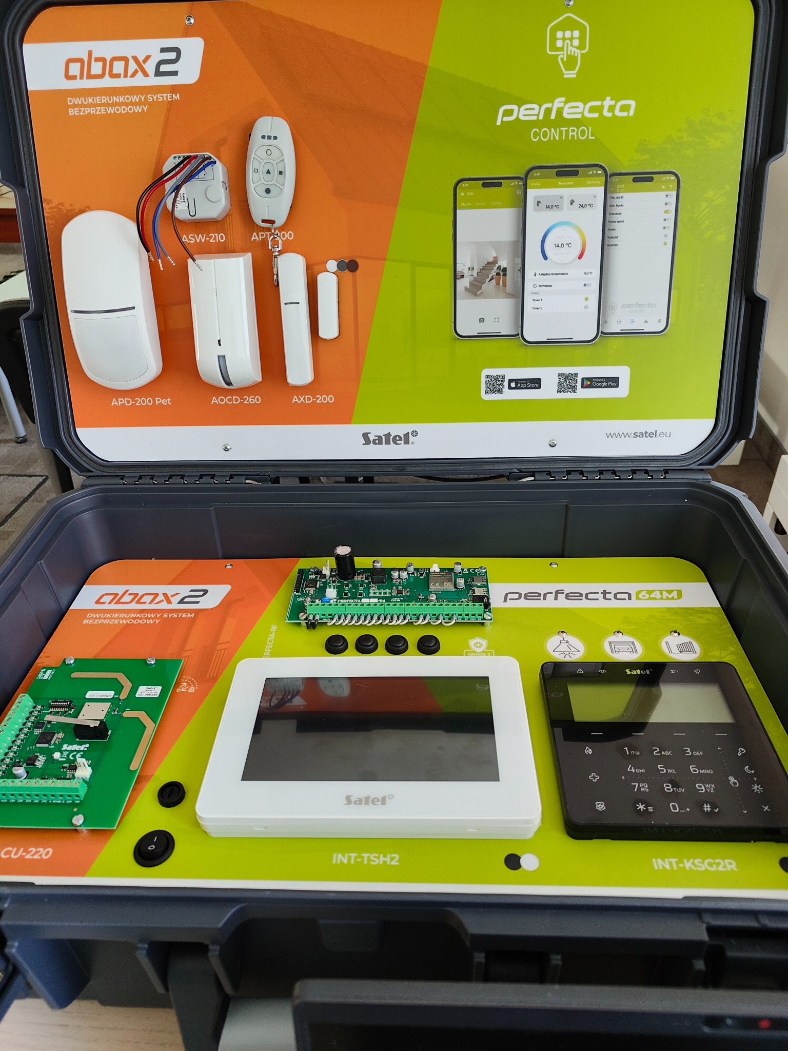 Zestaw demonstracyjny systemu alarmowego w otwartej walizce, prezentujący centralę, manipulatory, detektory i aplikację mobilną na smartfonach, marki Satel.
