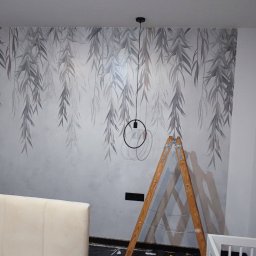 Lux Interiors usługi remontowo wykończeniowe - Ściana w pokoju z dekoracyjną tapetą w motywie liści wierzby i minimalistycznymi lampami wiszącymi, drewniana drabina oparta o ścianę, fragment łóżka z beżowym zagłówkiem i dziecięcego łóżeczka.