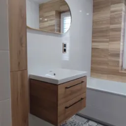 Nowoczesna łazienka z drewnianymi akcentami: szafka pod umywalkę z dwiema szufladami, owalne lustro z podświetleniem LED, płytki imitujące drewno na ścianie i wzorzysta podłoga.