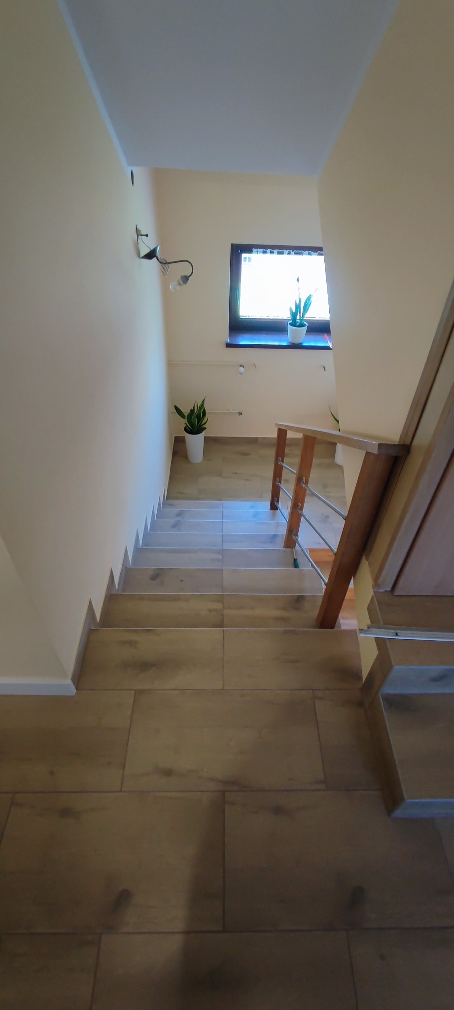 Schody wyłożone panelami imitującymi drewno, z drewnianą barierką z metalowymi elementami, oświetlone światłem z okna z roślinami i kinkietem na ścianie.