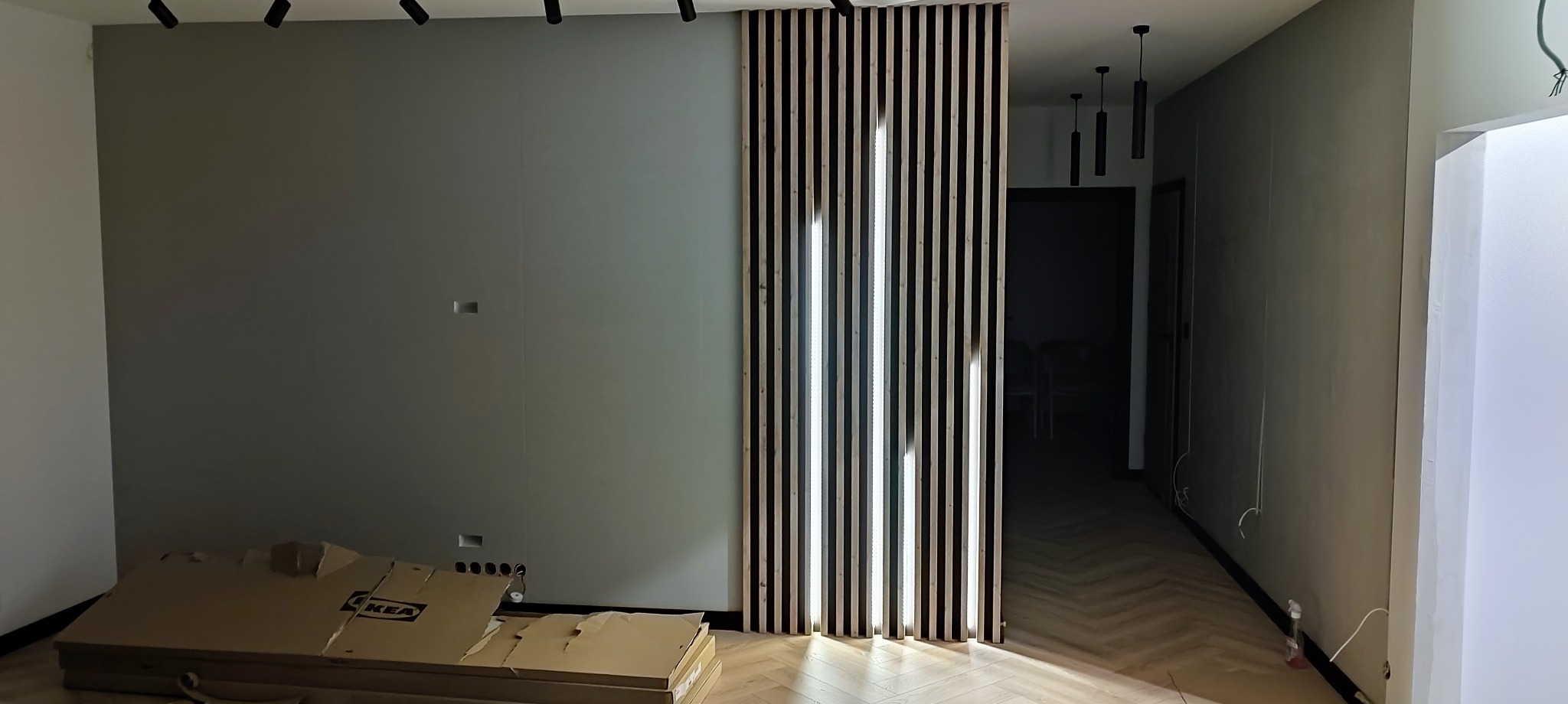 Szara ściana w trakcie prac wykończeniowych, widoczne kartony po meblach, drewniane lamele pionowe jako element dekoracyjny, oświetlenie szynowe na suficie.