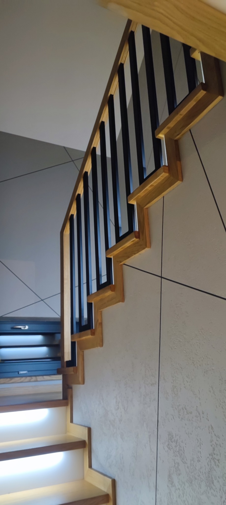 Nowoczesne schody z drewnianymi stopniami podświetlonymi LED i czarną balustradą na tle ściany z dekoracyjną fakturą.