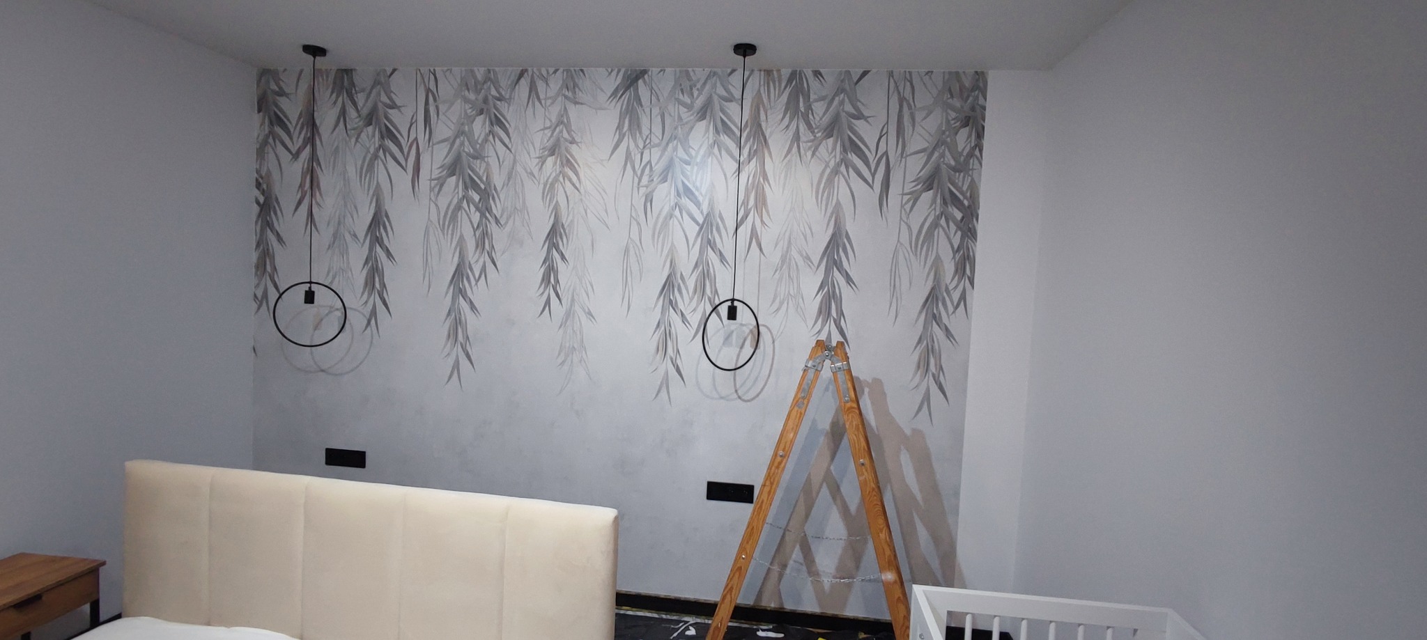 Ściana w pokoju z dekoracyjną tapetą w motywie liści wierzby i minimalistycznymi lampami wiszącymi, drewniana drabina oparta o ścianę, fragment łóżka z beżowym zagłówkiem i dziecięcego łóżeczka.