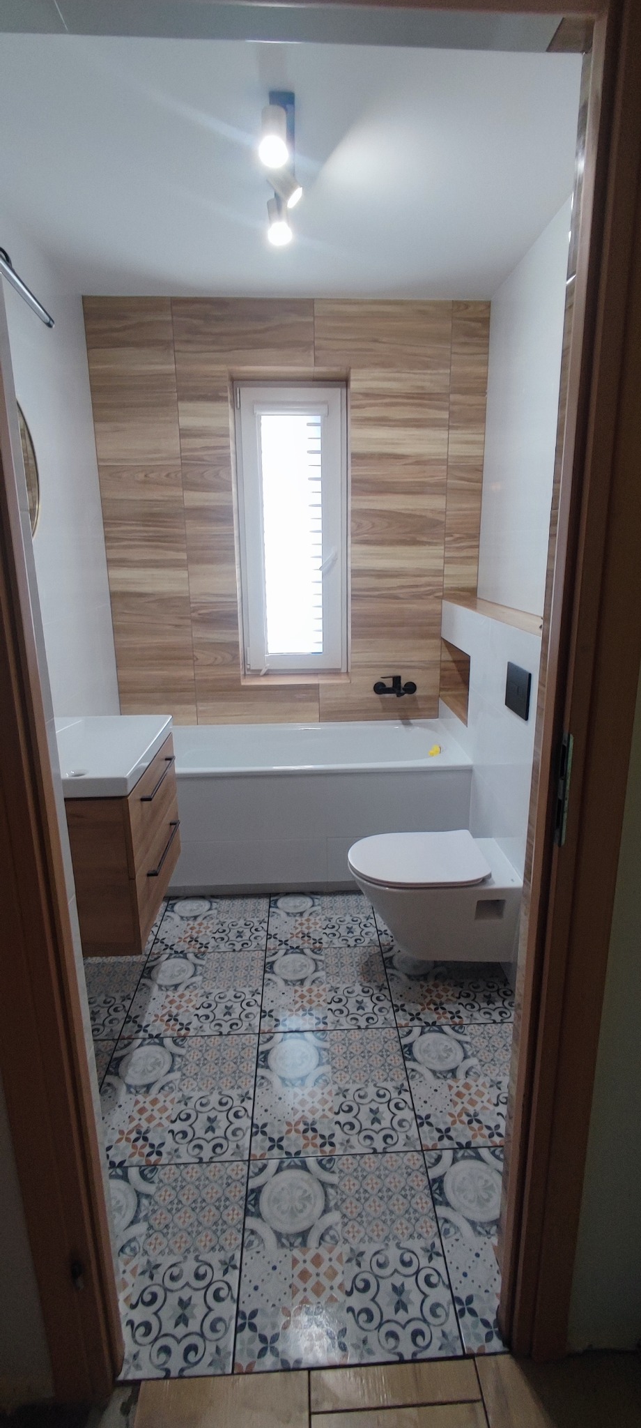 Ukończona łazienka z drewnopodobnymi płytkami ściennymi, wzorzystą podłogą, białą wanną i podwieszaną toaletą, widziana zza framugi drzwi.