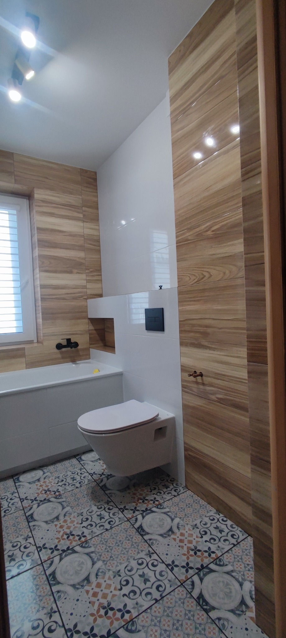 Nowoczesna łazienka po remoncie: drewnopodobne płytki na ścianach, wzorzysta podłoga, podwieszana toaleta i biała wanna z czarną armaturą.