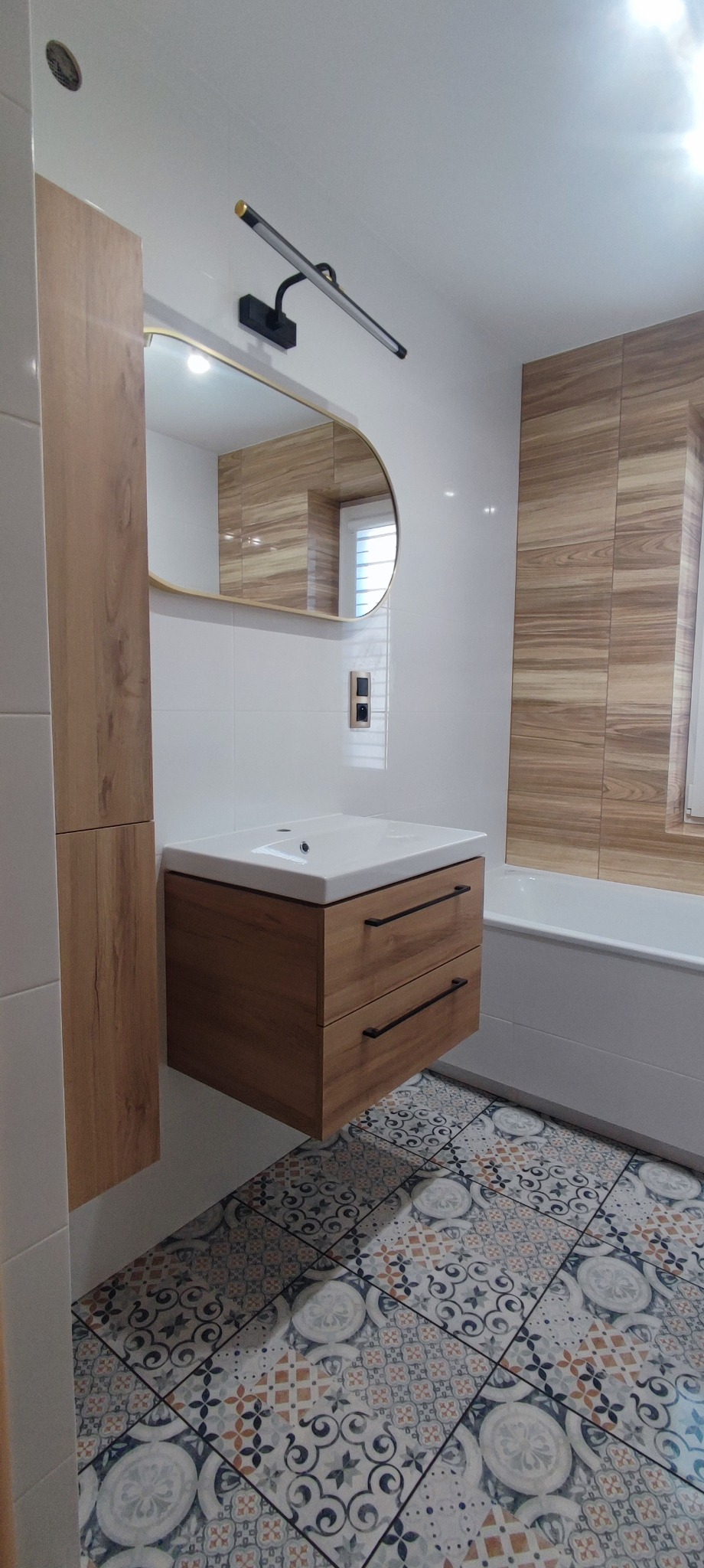 Nowoczesna łazienka z drewnianymi akcentami: szafka pod umywalkę z dwiema szufladami, owalne lustro z podświetleniem LED, płytki imitujące drewno na ścianie i wzorzysta podłoga.