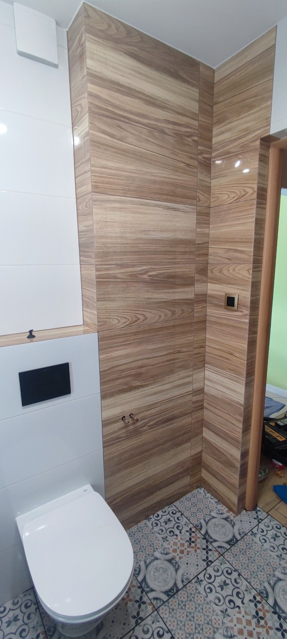 Nowoczesna toaleta podwieszana z białą ceramiką i zabudową, ściany wyłożone płytkami imitującymi drewno z widocznymi zaworami, podłoga w patchworkową mozaikę.