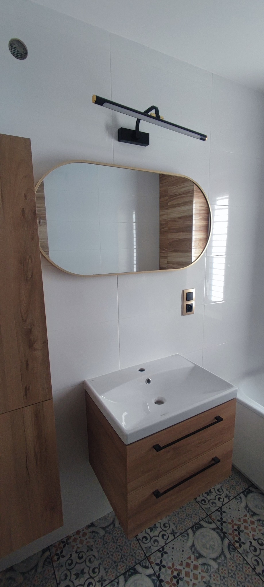 Nowoczesna łazienka z owalnym lustrem z podświetleniem LED, białą umywalką nablatową na drewnianej szafce z czarnymi uchwytami, oraz wzorzystą podłogą z płytek.