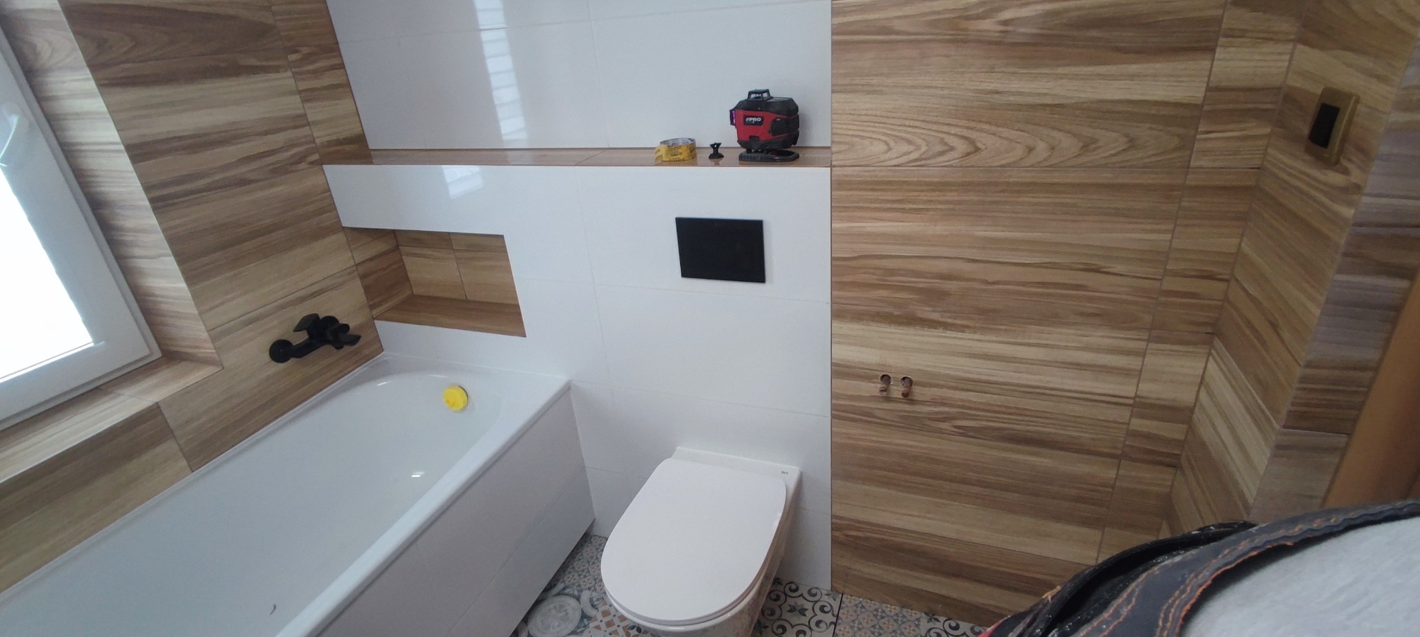 Nowoczesna łazienka z drewnopodobnymi płytkami, białą armaturą i czarnymi akcentami, widoczny poziomica laserowa na półce.