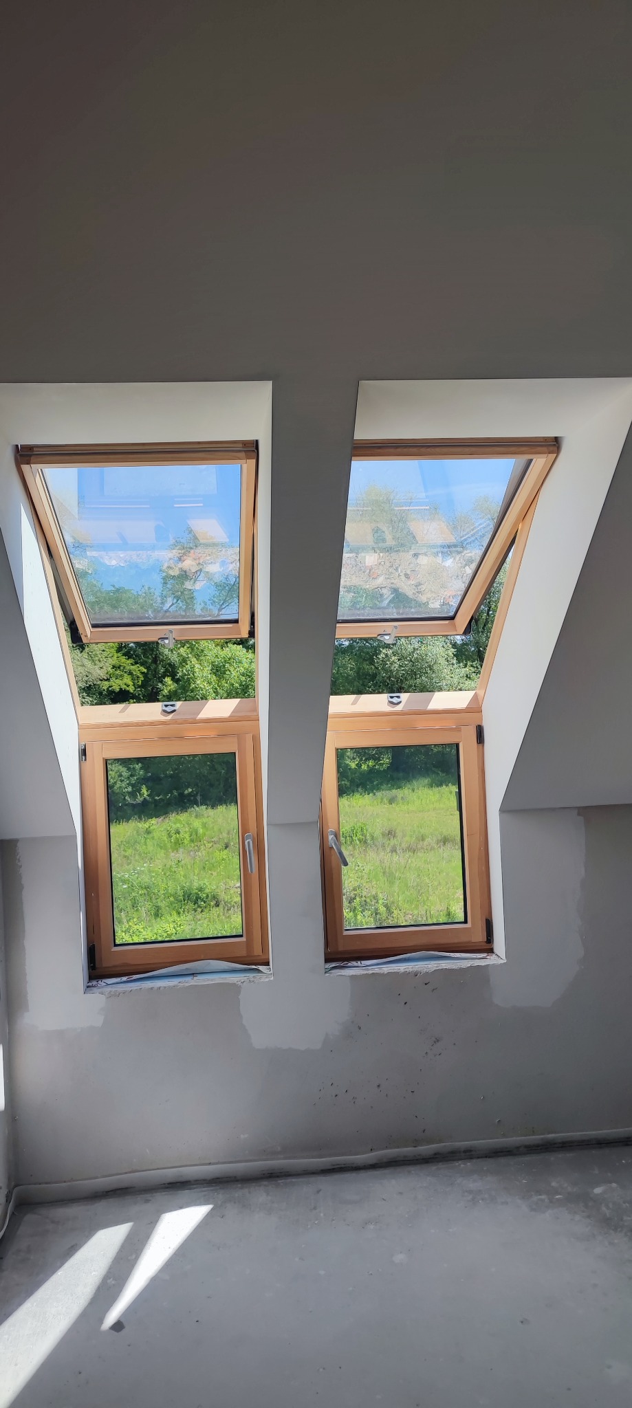 Dwa otwarte okna dachowe z drewnianymi ramami, widok na zieleń zewnątrz, ściany w trakcie wykończenia.