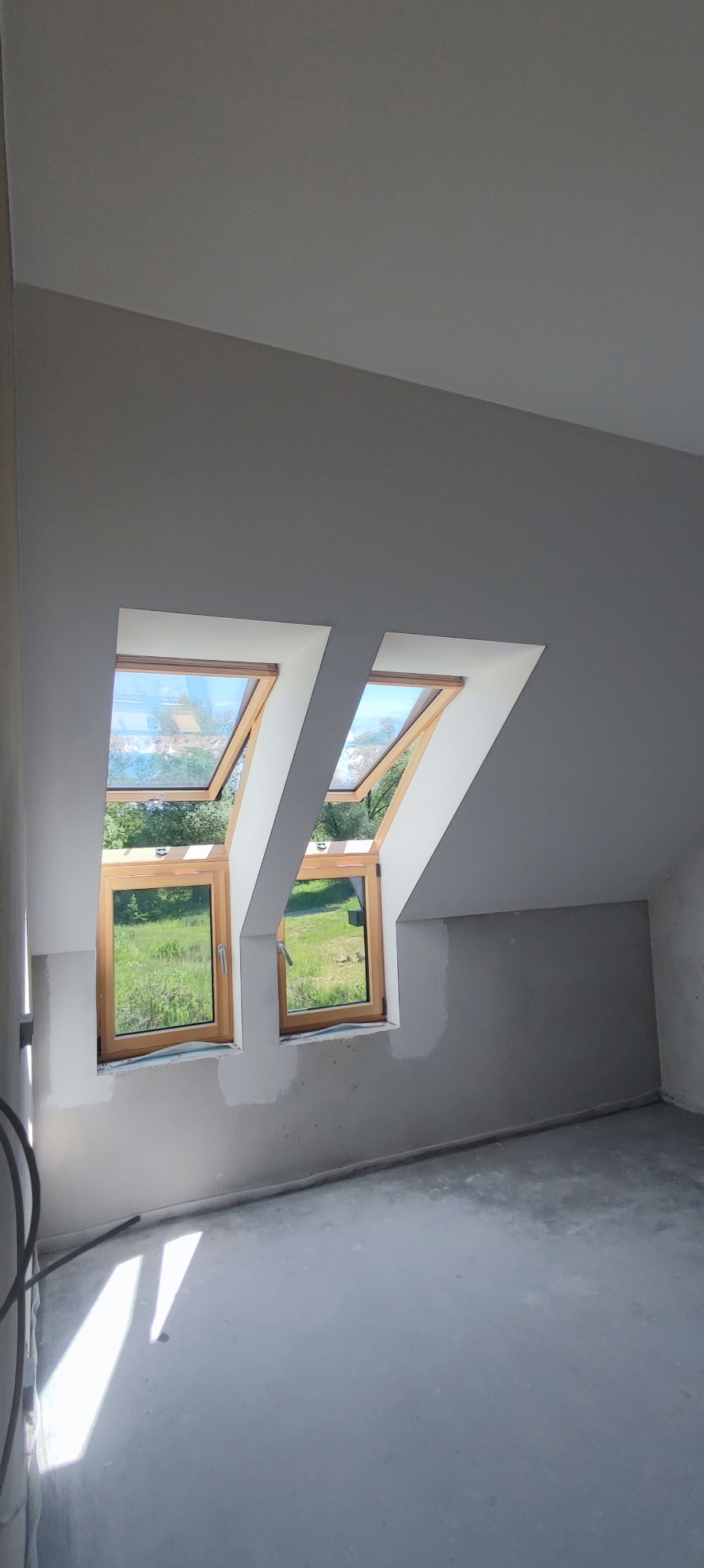 Poddasze w trakcie prac wykończeniowych: dwa drewniane okna dachowe, ściany w szarym kolorze po położeniu gładzi, surowa podłoga, widoczne ślady po pracach malarskich.