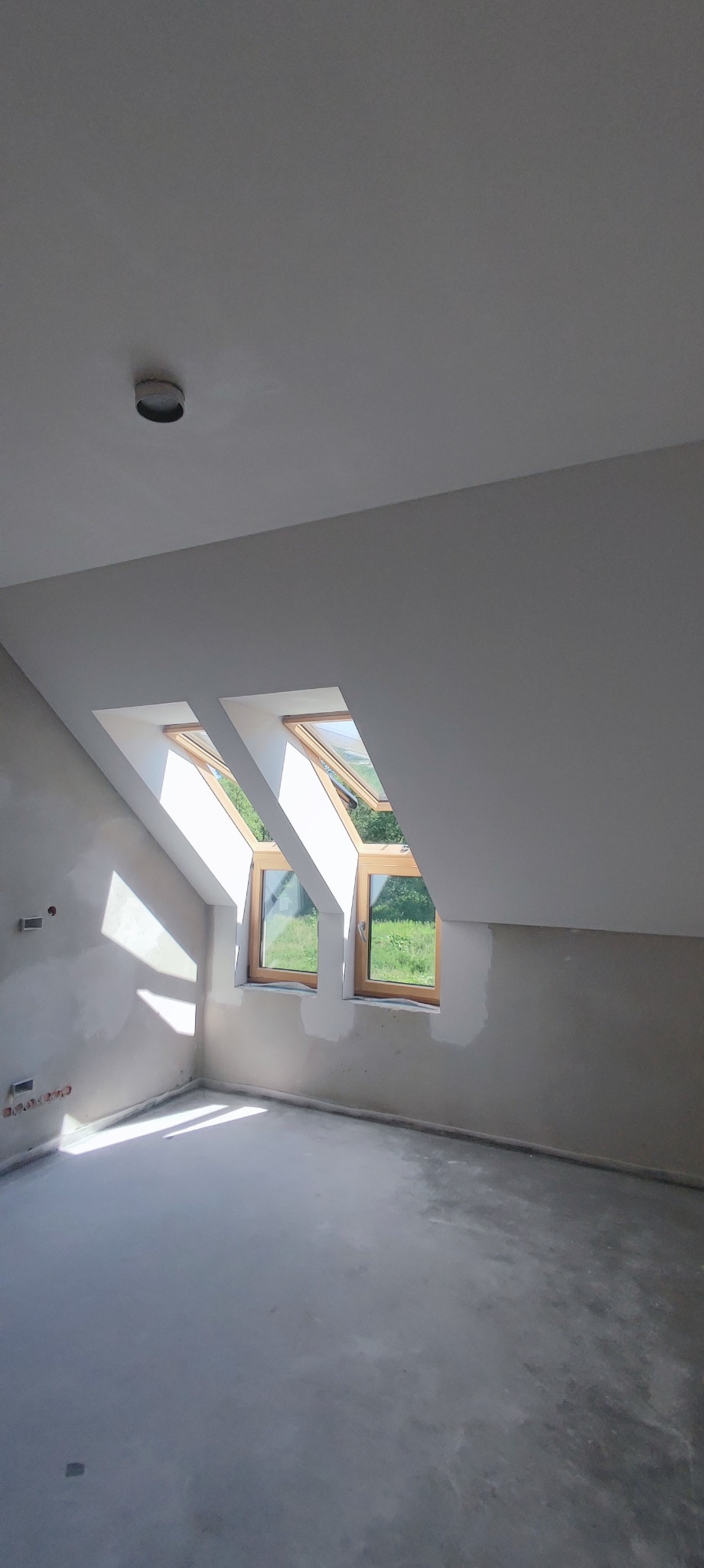 Pokój na poddaszu w trakcie prac wykończeniowych, widoczne dwa okna dachowe, ściany częściowo zagruntowane, podłoga wylewka, surowy stan wykończenia.