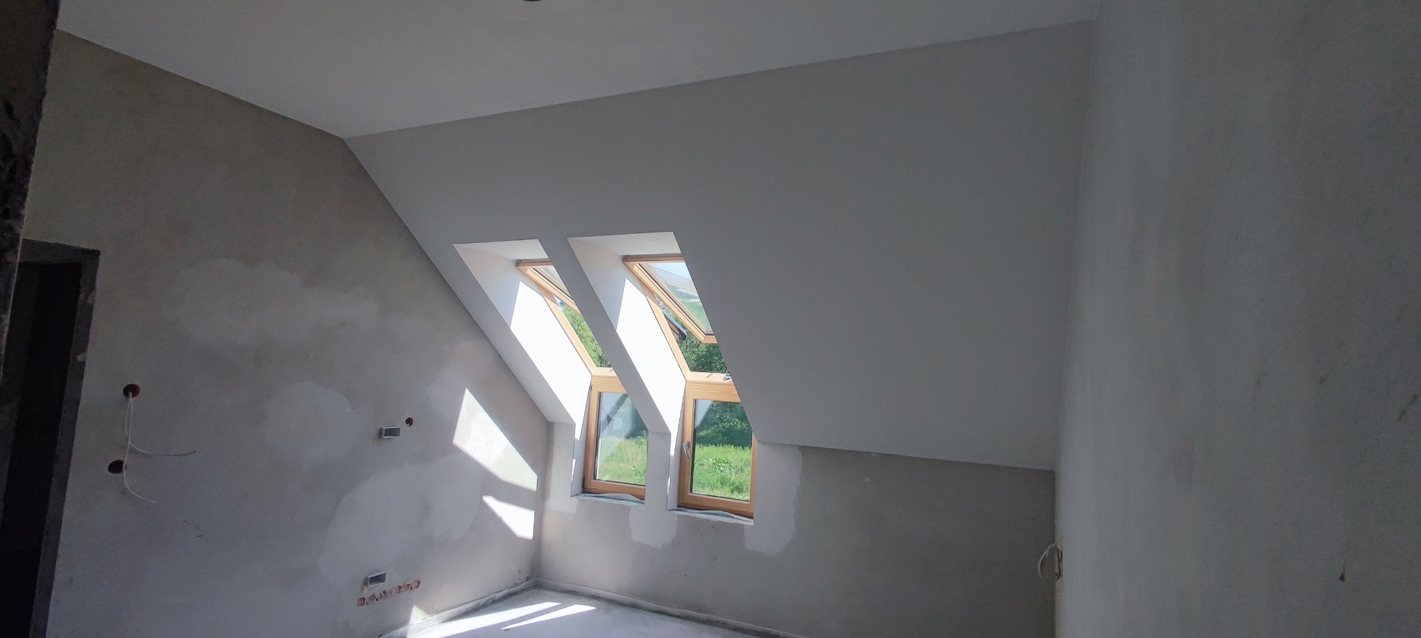 Pokój na poddaszu w trakcie prac wykończeniowych: ściany częściowo otynkowane, dwa okna dachowe z widokiem na zieleń.