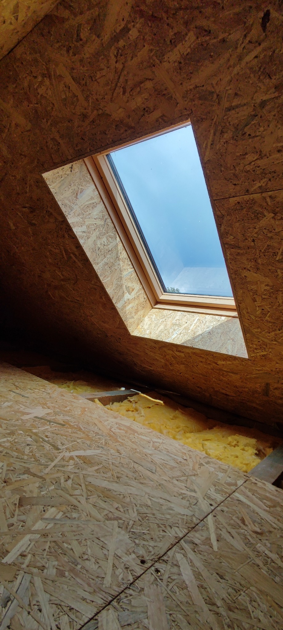 Wnętrze poddasza z oknem dachowym, widoczne płyty OSB i izolacja z wełny mineralnej, wpadające światło dzienne.