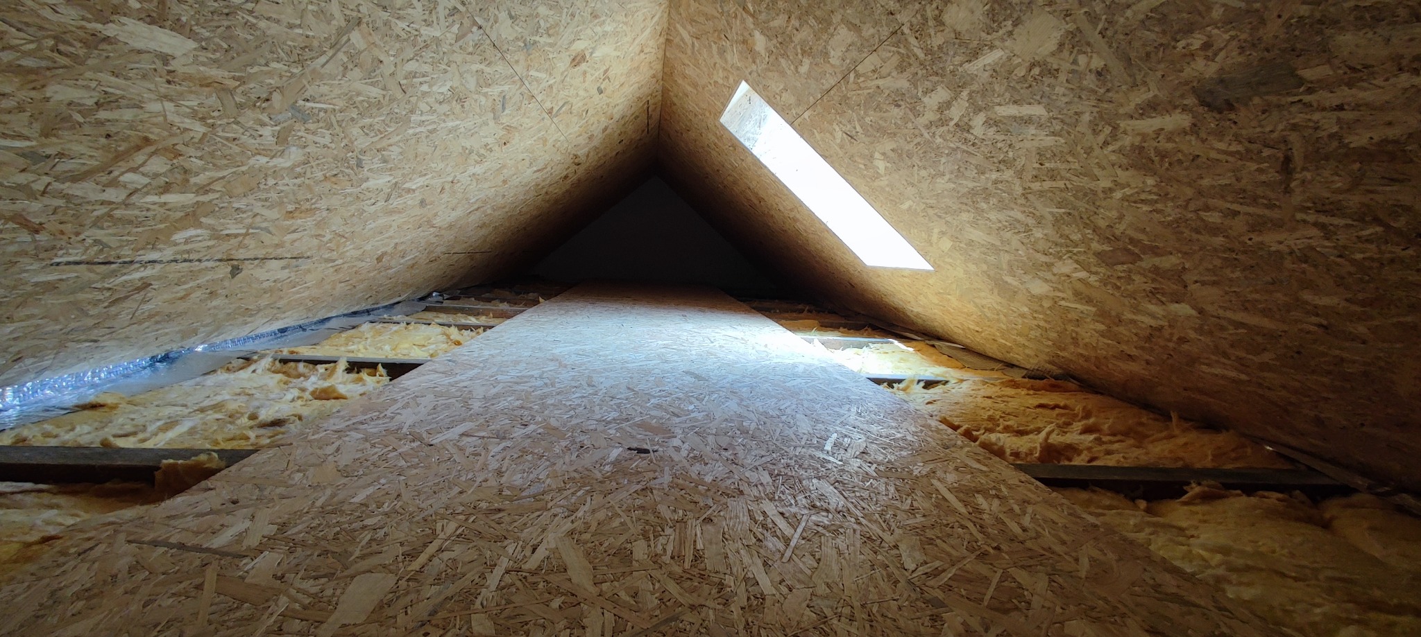Poddasze z widoczną izolacją z wełny mineralnej i płytami OSB, oświetlone przez okno dachowe.