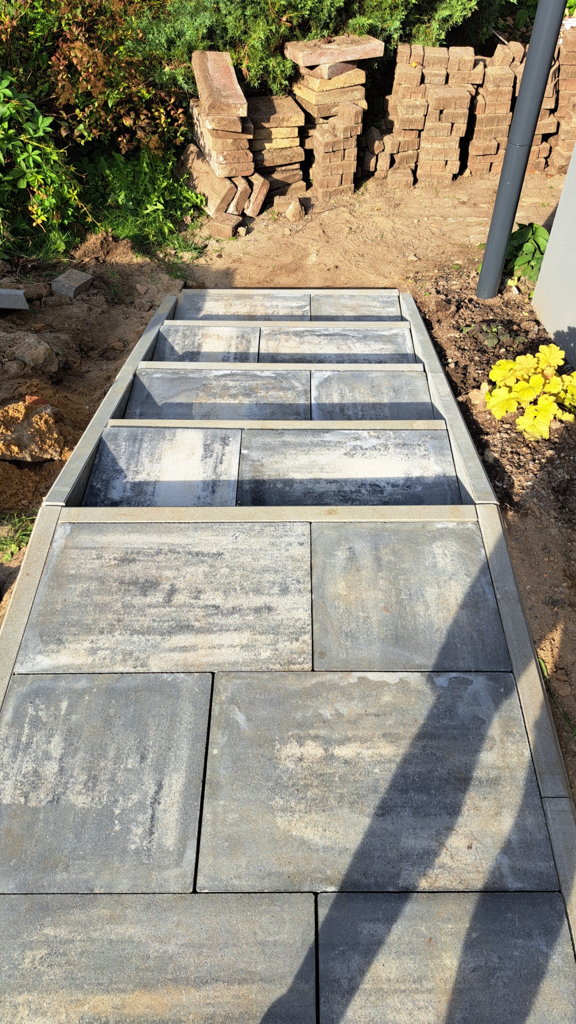 Schody z szarych, prostokątnych płyt betonowych, obramowane metalową listwą, prowadzące w górę, z fragmentami kostki brukowej obok i roślinnością w tle.