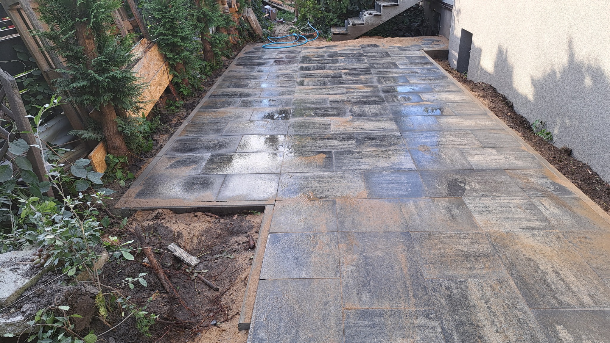 Świeżo ułożona, mokra nawierzchnia z kostki w odcieniach szarości i brązu, z widocznymi śladami piasku i wody, otoczona zielenią i elementami konstrukcyjnymi.